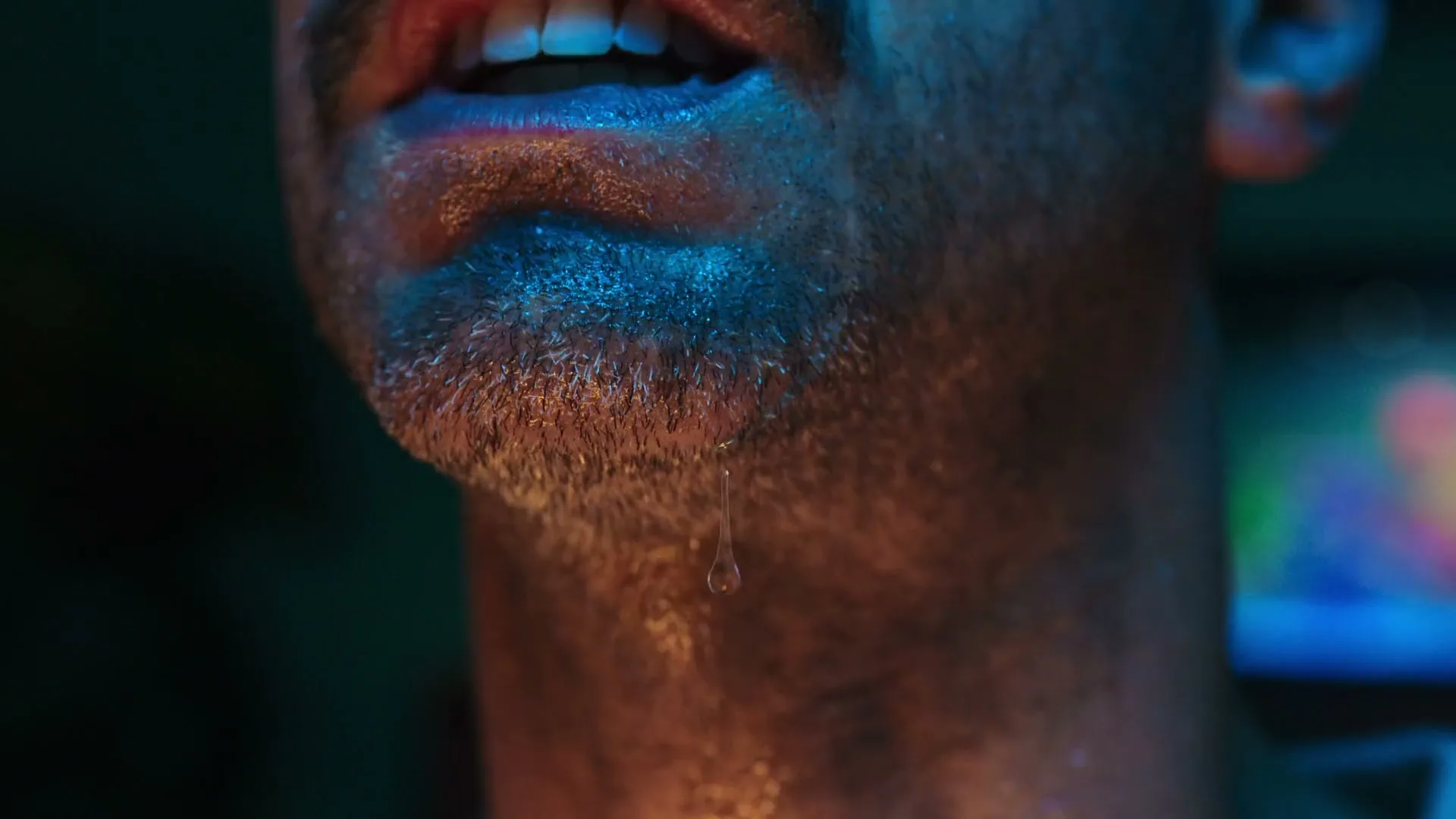A man's chin and mouth are shown in a closeup, with sweat dripping from his chin, in a dark interior setting.