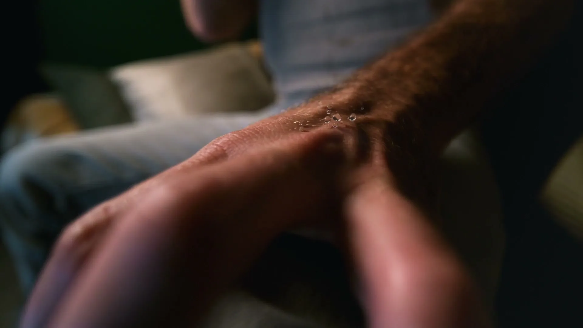 A man's arm with goosebumps and sweat is shown, then a drop of sweat reflects a man's face, in a dark room.