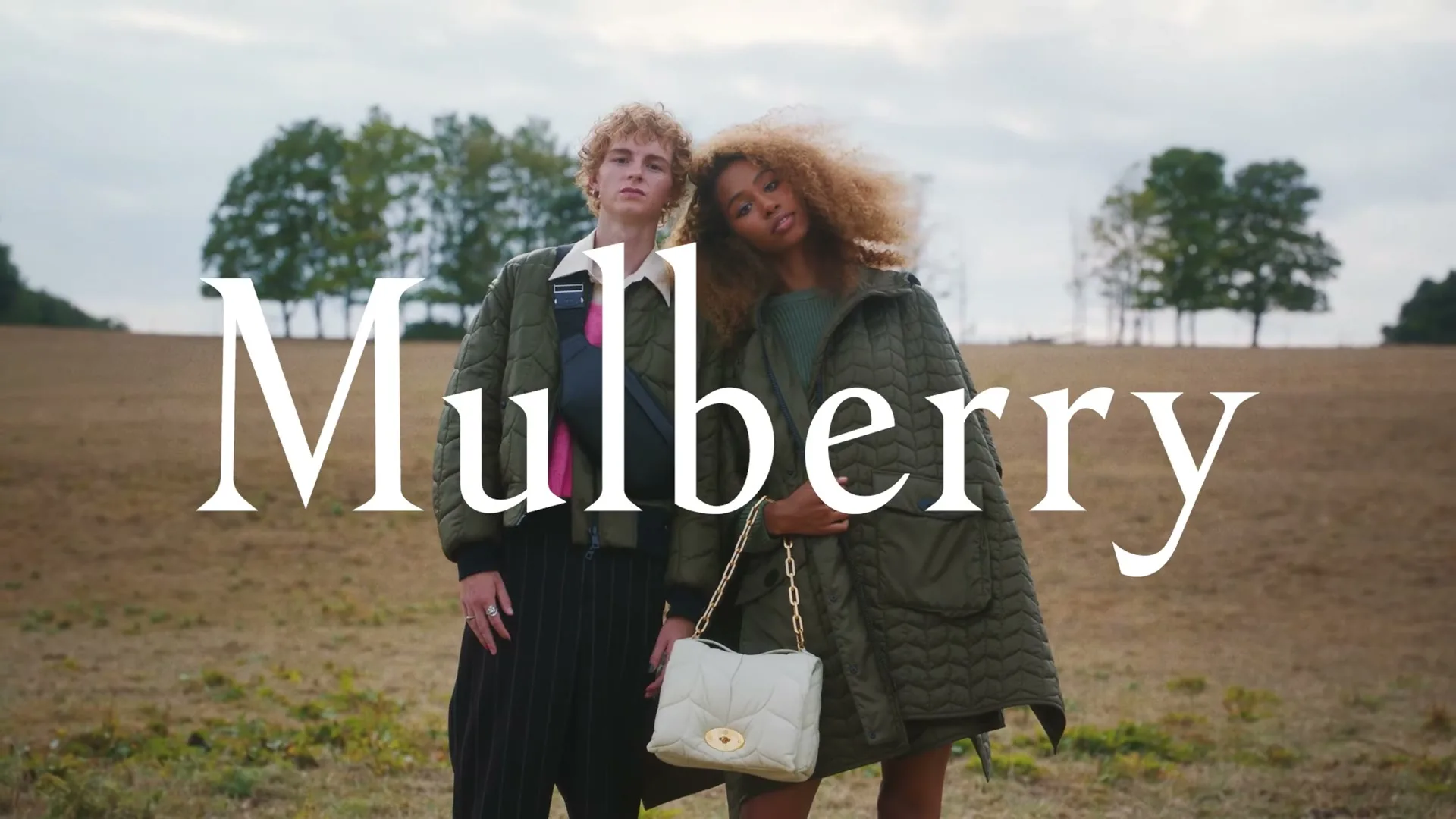 A young man and woman stand in a field, wearing quilted jackets, with the brand name "Mulberry" superimposed over them.