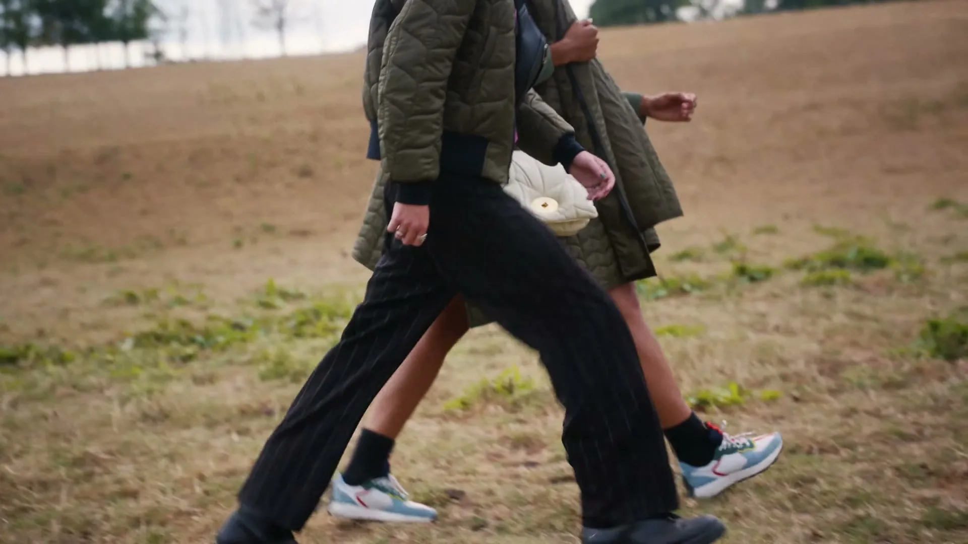 Two people, one in a green quilted jacket and black pants, the other in a green quilted jacket and skirt, walk across a grassy field.