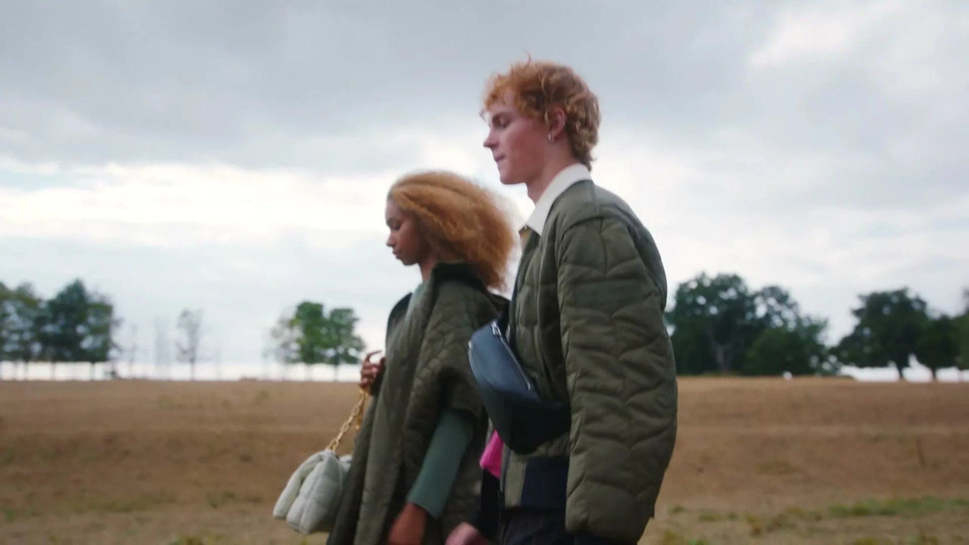 A young man and woman walk side-by-side across a dry, grassy field under a cloudy sky.