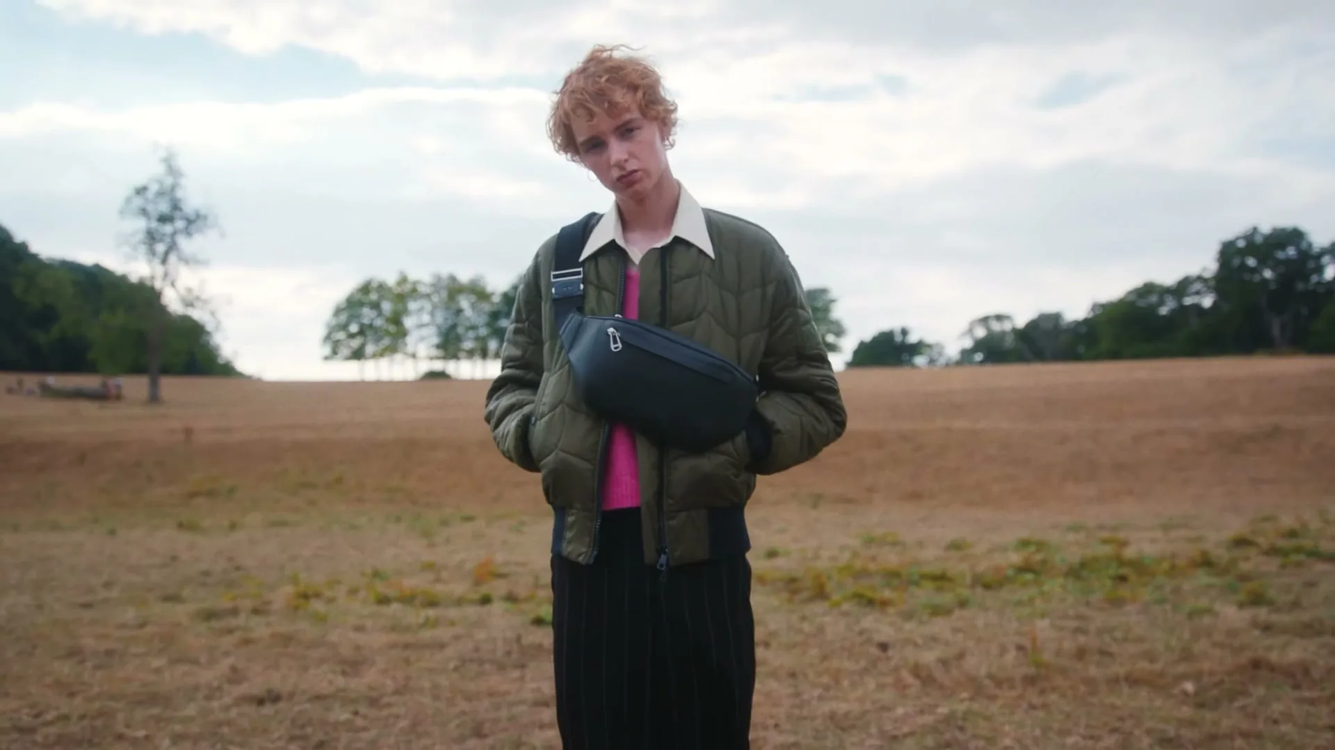 A young man with curly red hair stands in a dry field, wearing a green quilted jacket and a black fanny pack.