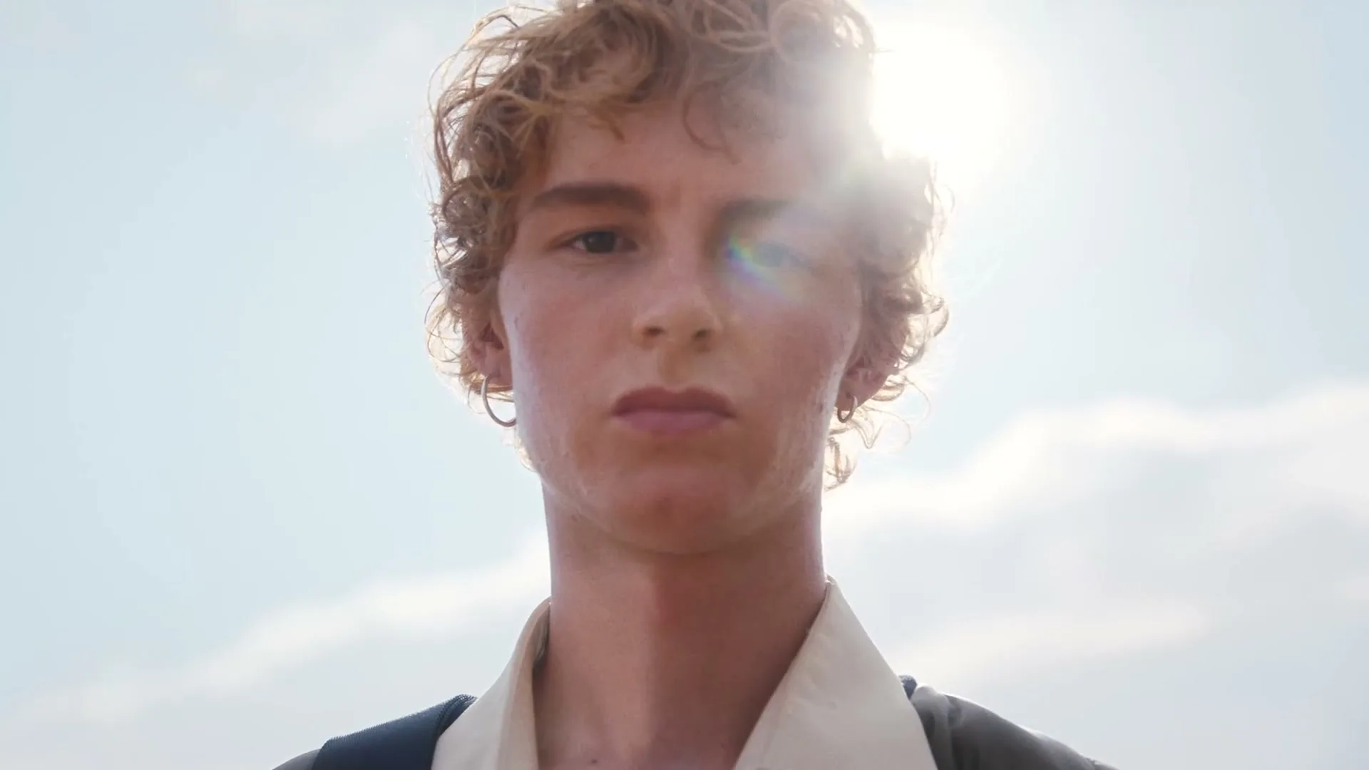 A young man with curly red hair looks directly at the camera, with the sun flaring behind his head against a bright sky.