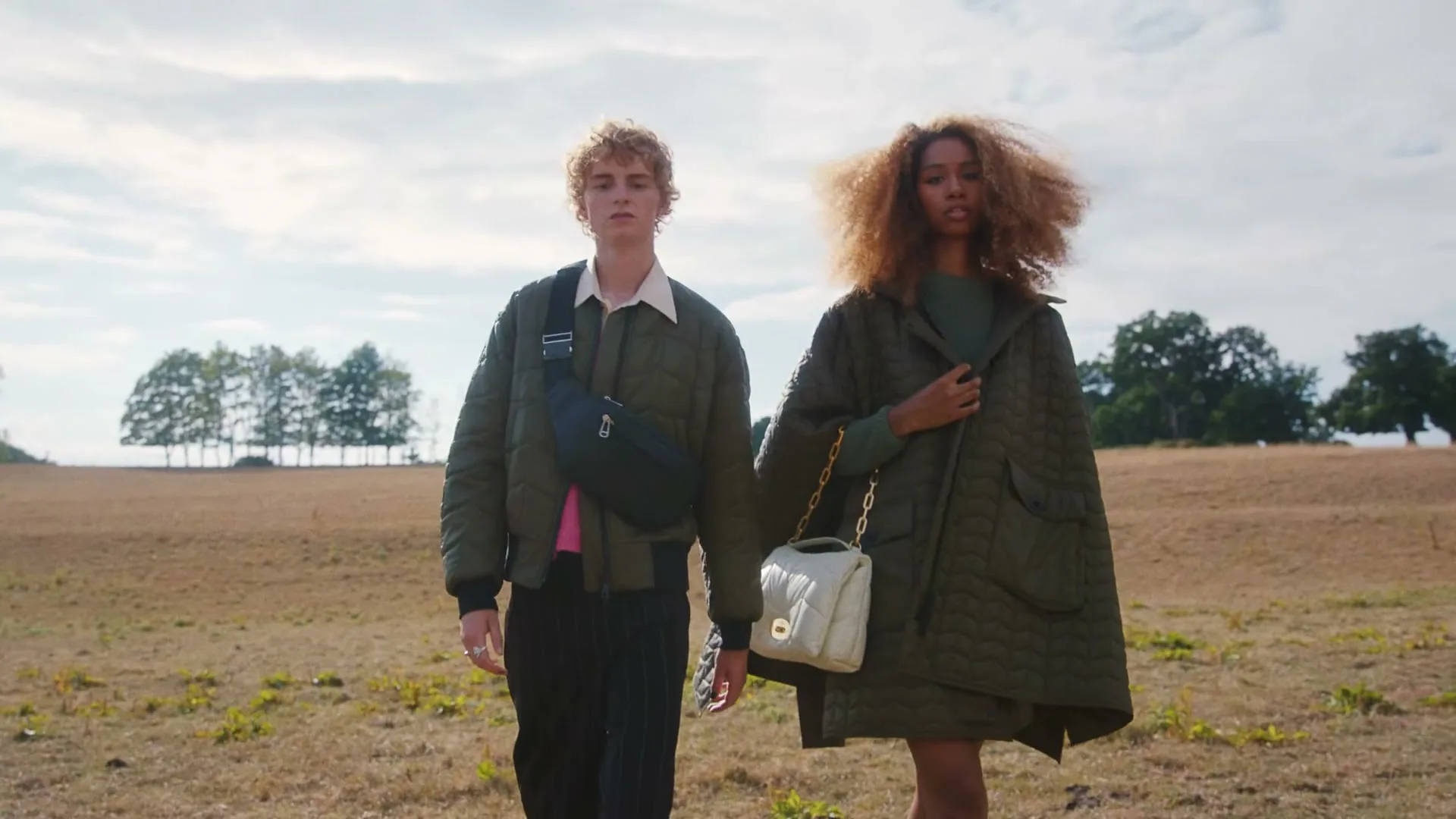 A young man and woman, both wearing green quilted jackets and carrying bags, walk towards the camera in a dry, grassy field under a bright sky.