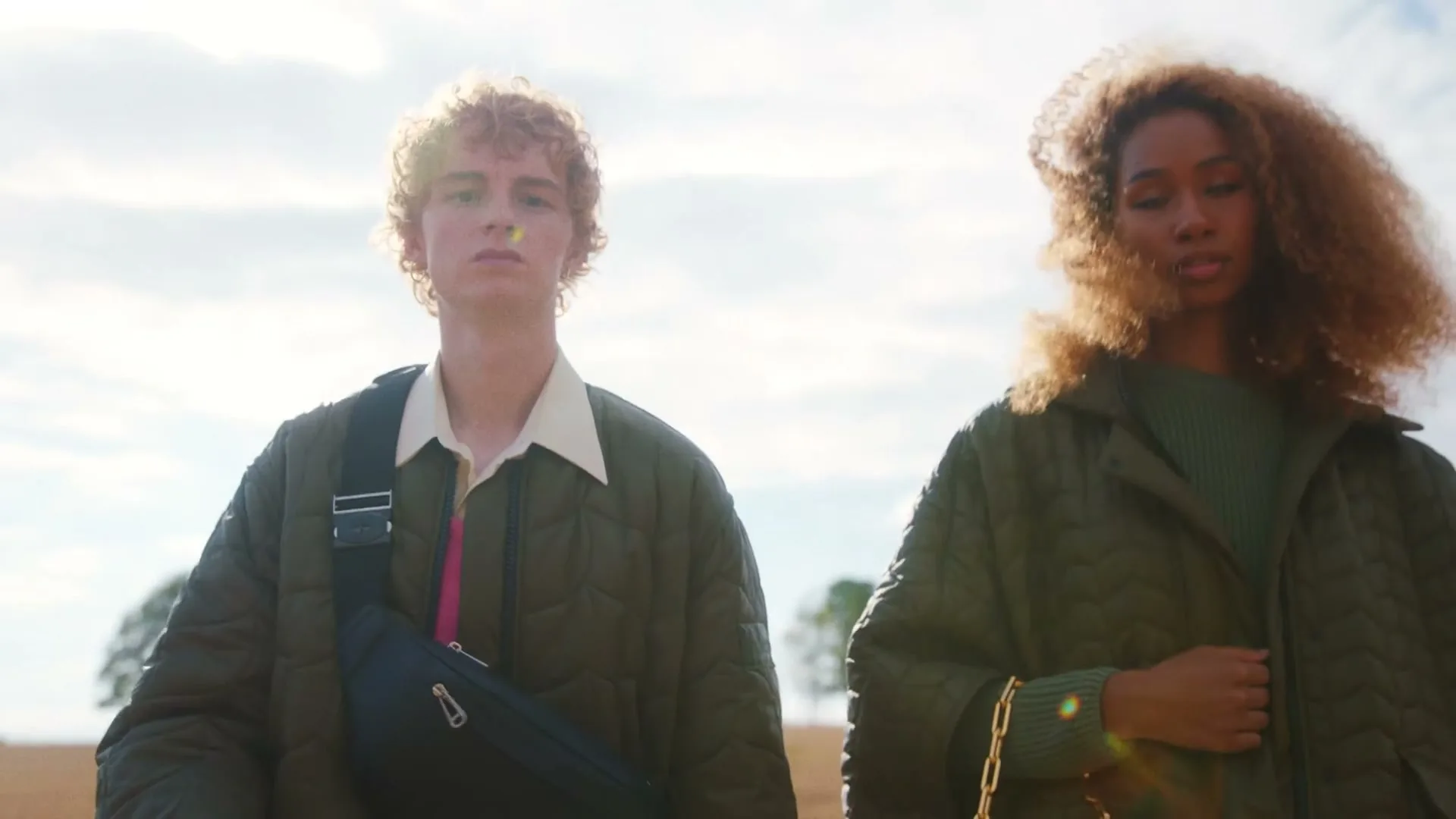 A young man and woman, both wearing green quilted jackets, stand side-by-side in a field under a bright sky, looking directly at the camera.