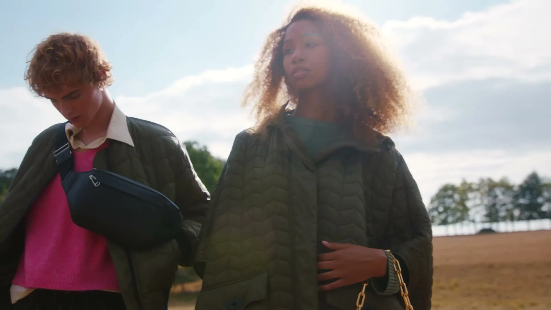 A young man and woman walk side-by-side in quilted jackets and a field under a bright sky.