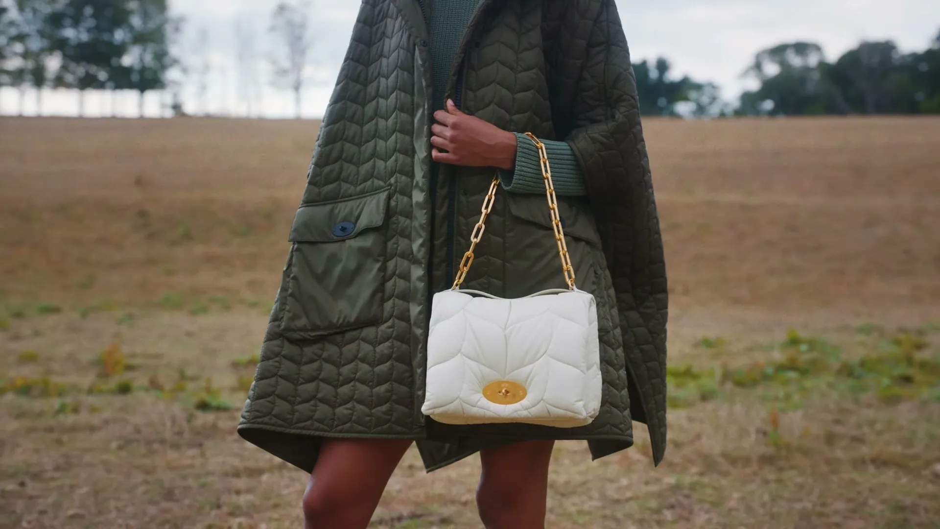 A person in a green quilted coat holds a white quilted handbag with a gold chain strap in a field.
