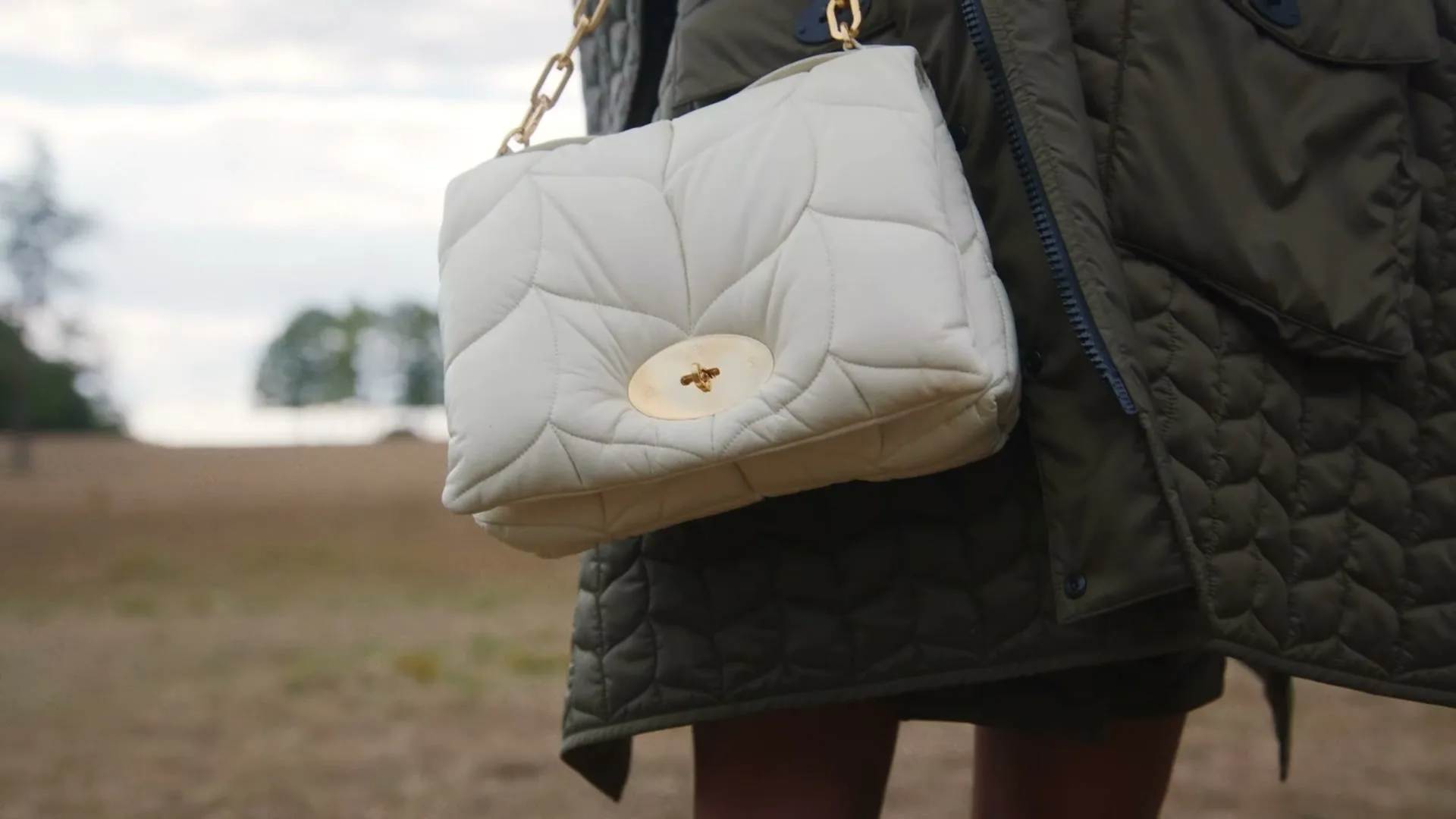 A person wearing a green quilted jacket holds a white quilted handbag with a gold chain strap, standing in an open field under a cloudy sky.