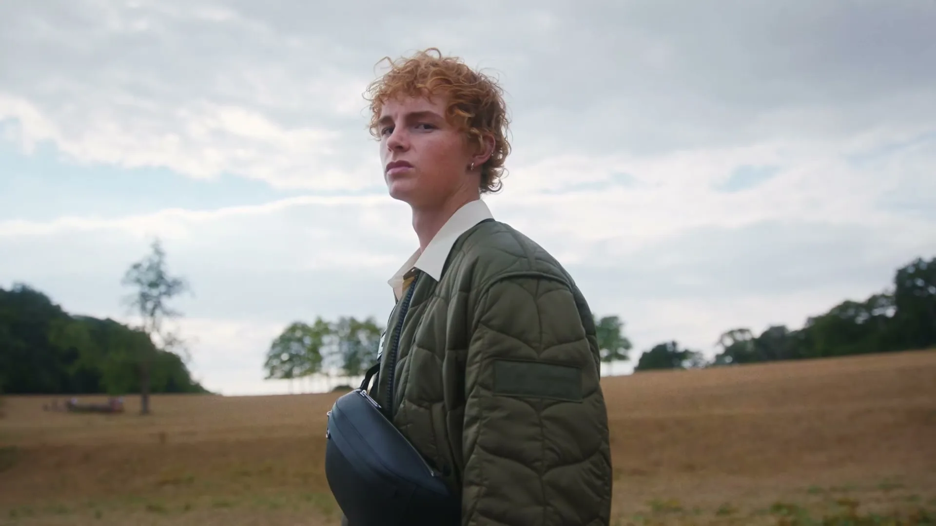 A young man with red curly hair, wearing a green quilted jacket and carrying a black bag, stands in a field and turns to look at the camera.