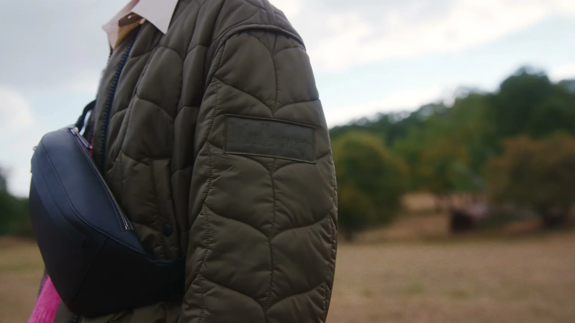 A person wearing a quilted jacket and a black fanny pack stands in a field with trees in the background.