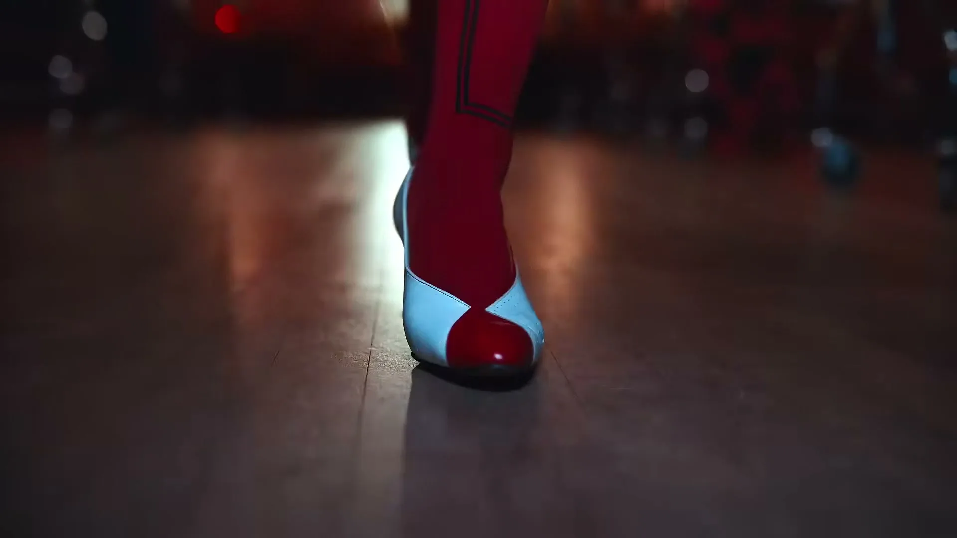 A person's legs and feet are shown walking on a wooden floor, wearing various stylish shoes, in a dimly lit interior.