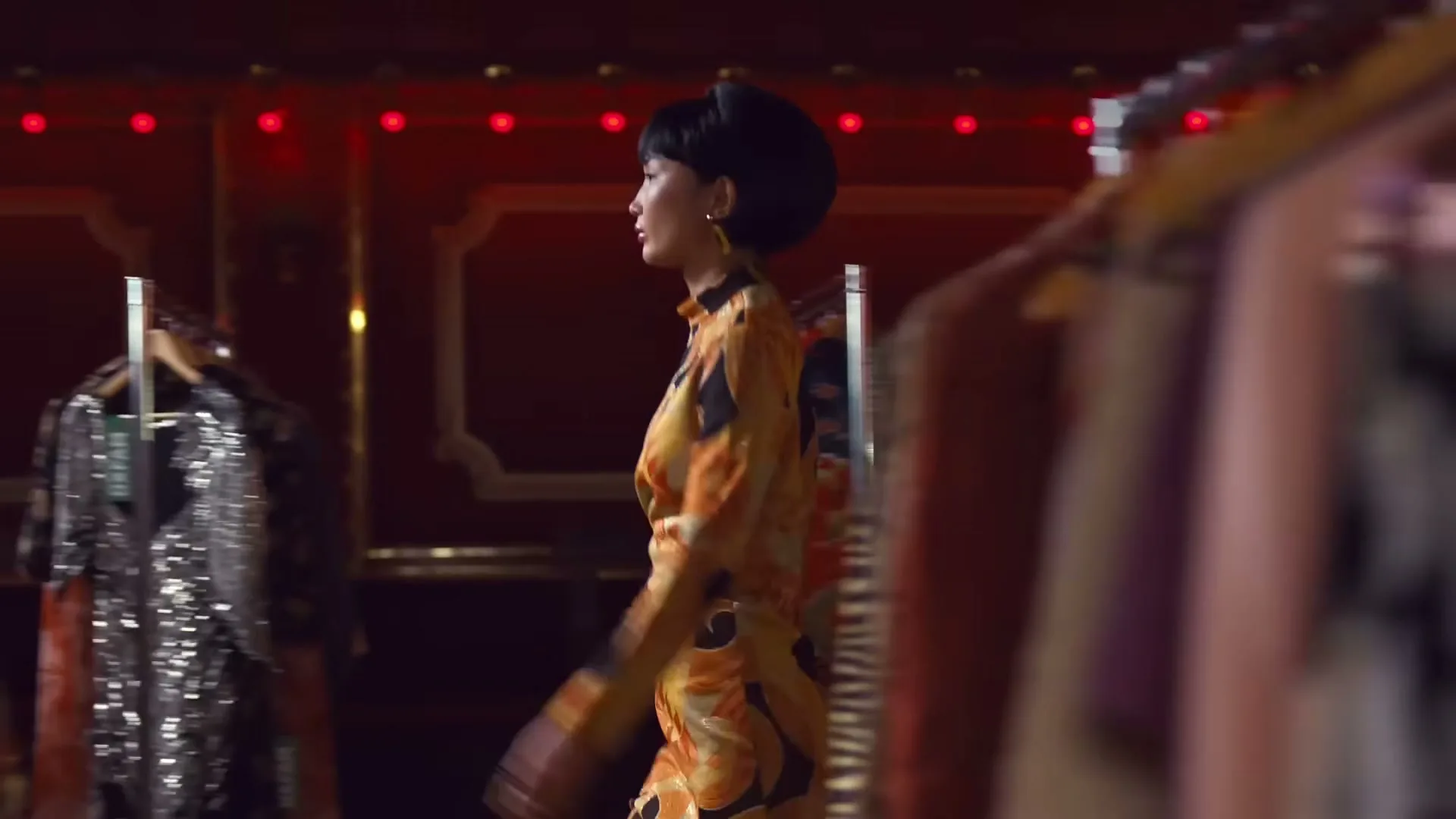 A woman with a bob haircut and a patterned dress walks through a vintage clothing store, looking at racks of clothes.