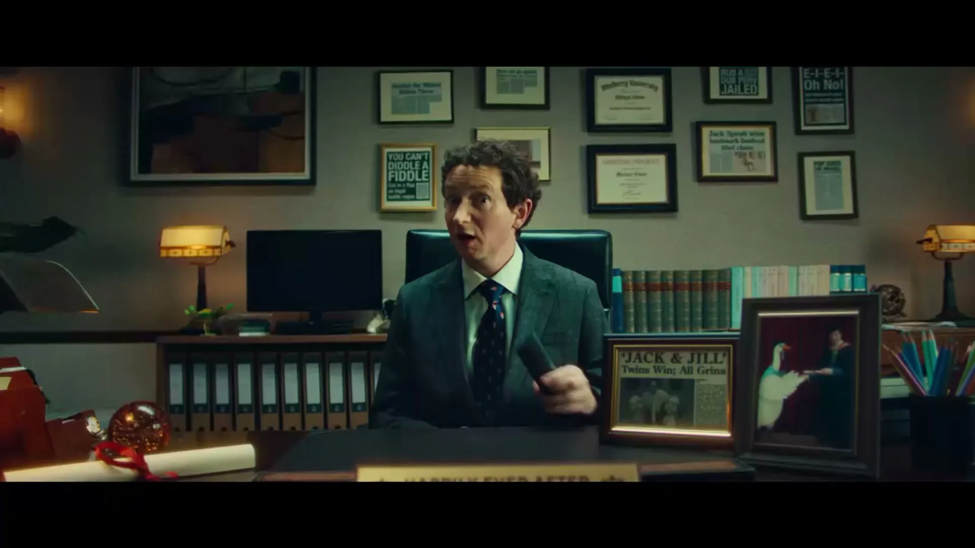 A male lawyer in a suit sits at his desk, talking and then turning to watch a TV commercial on a monitor in his office.