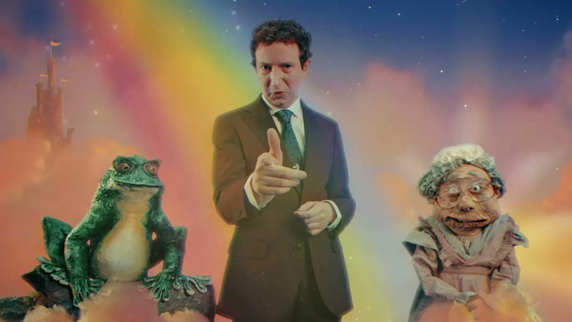 A man in a suit points at the camera, flanked by a frog and an old woman puppet, against a whimsical sky background with a castle.