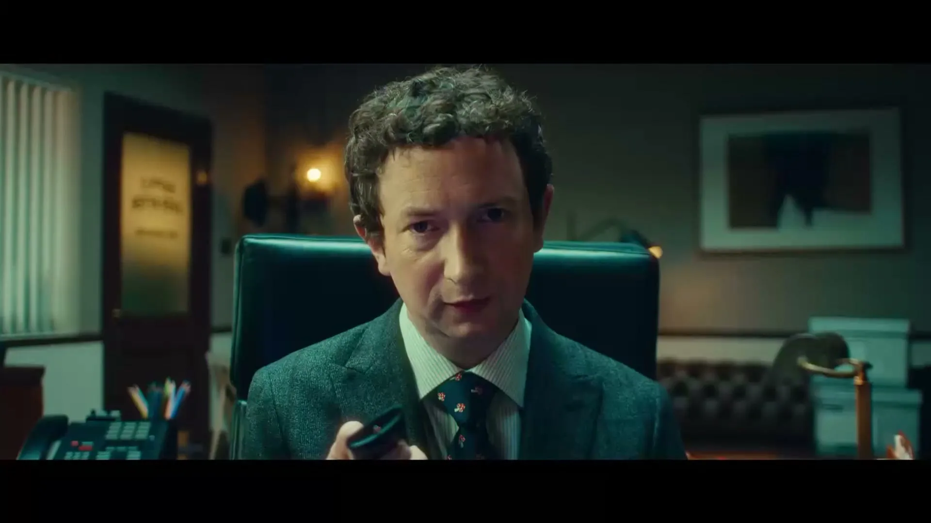 A man with curly hair and a suit sits at a desk, holding a small black object and speaking directly to the camera in an office.