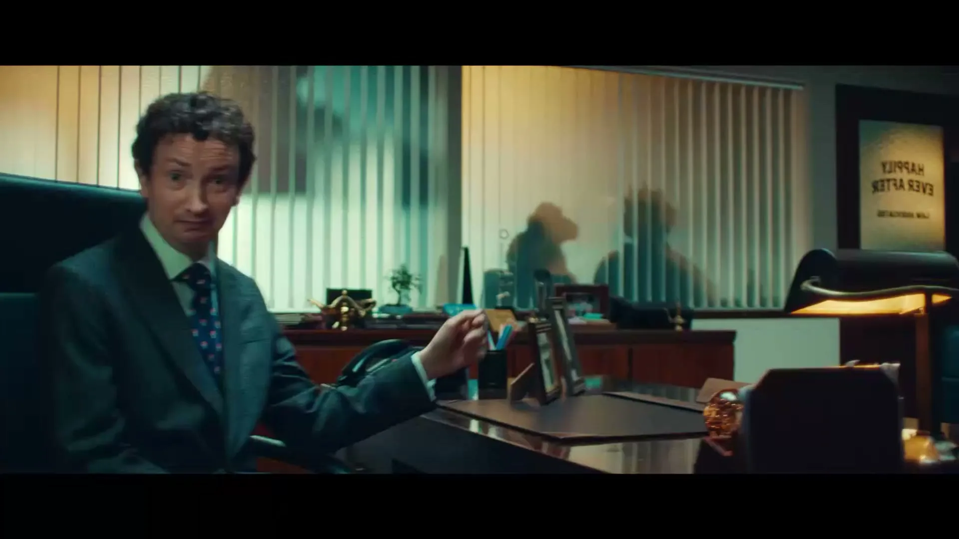 A male lawyer in a suit sits at his desk, talking to the camera, then slides out of frame as the camera pans right to show his office door.