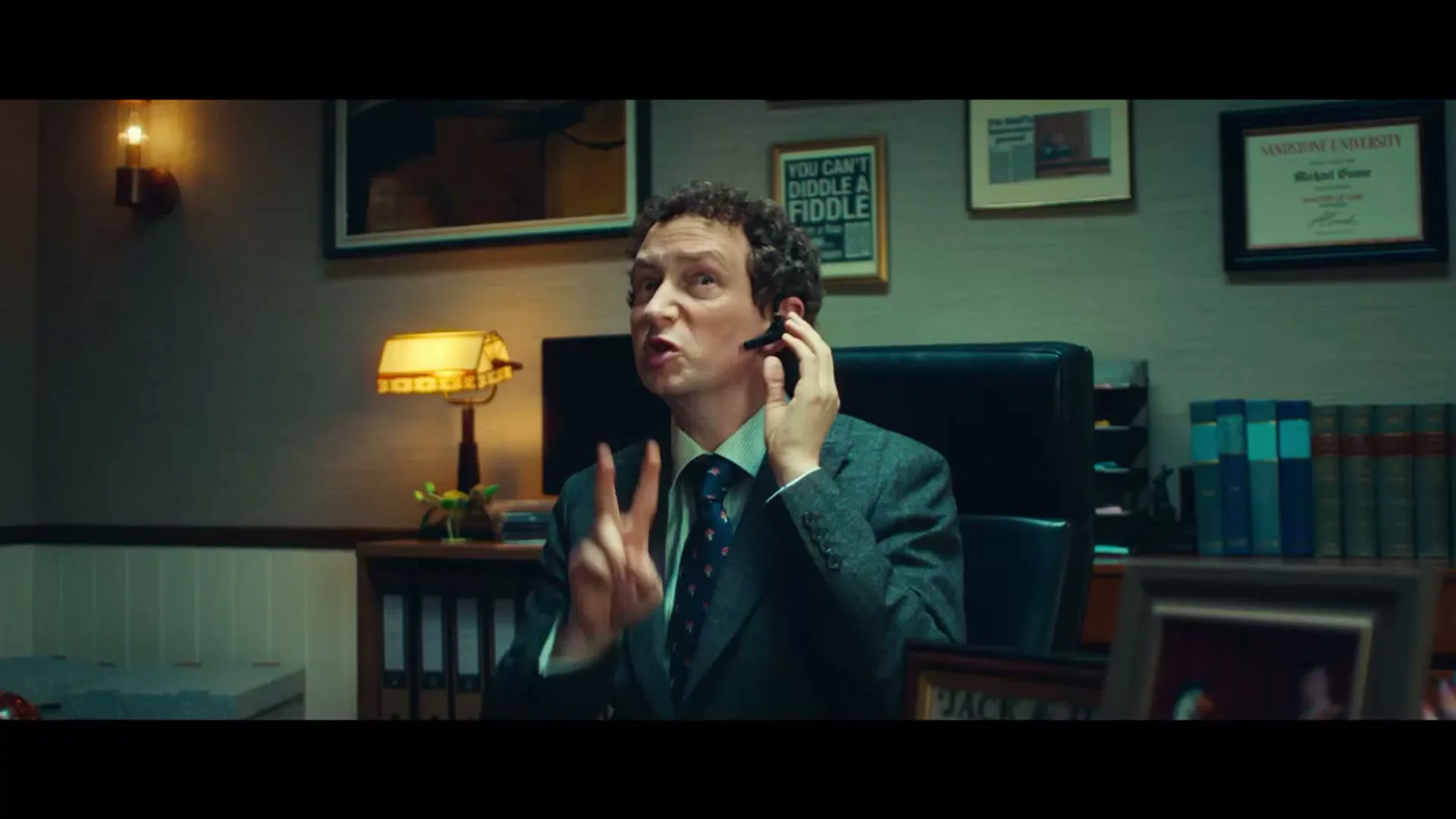 A man in a suit and tie, wearing a headset, sits at a desk in an office, talking and gesturing with his hand.