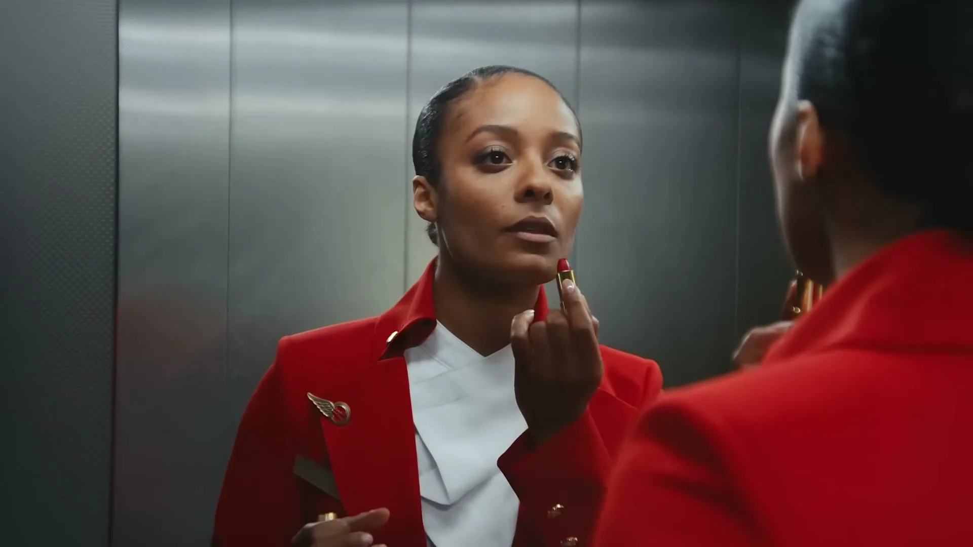 A female flight attendant in a red uniform applies lipstick while looking at her reflection in an elevator mirror, preparing for her shift.