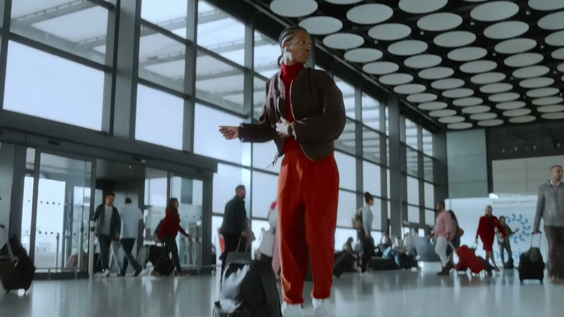 A young Black man in a brown jacket and red pants dances through a busy airport terminal, wearing wireless earbuds.