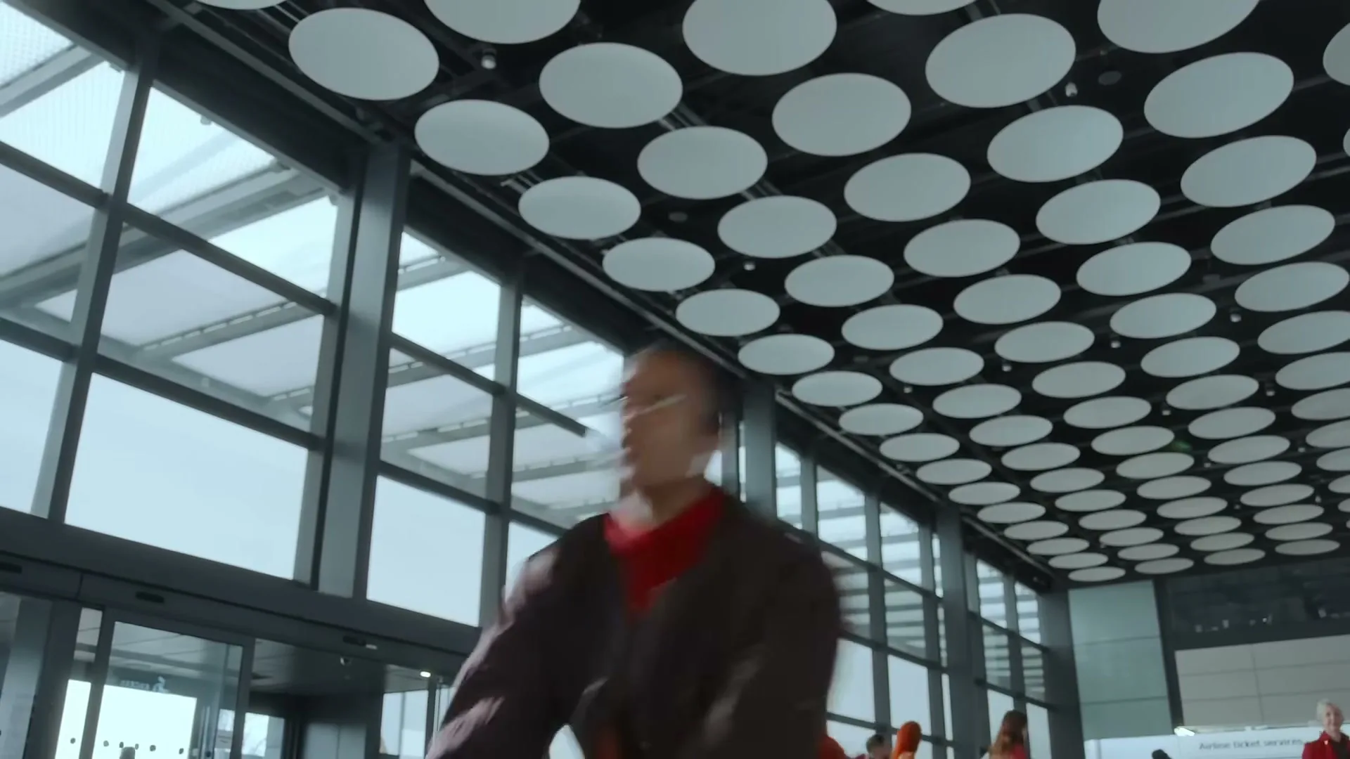 A man in a brown jacket and orange pants dances energetically in a modern airport terminal with large windows and a patterned ceiling.
