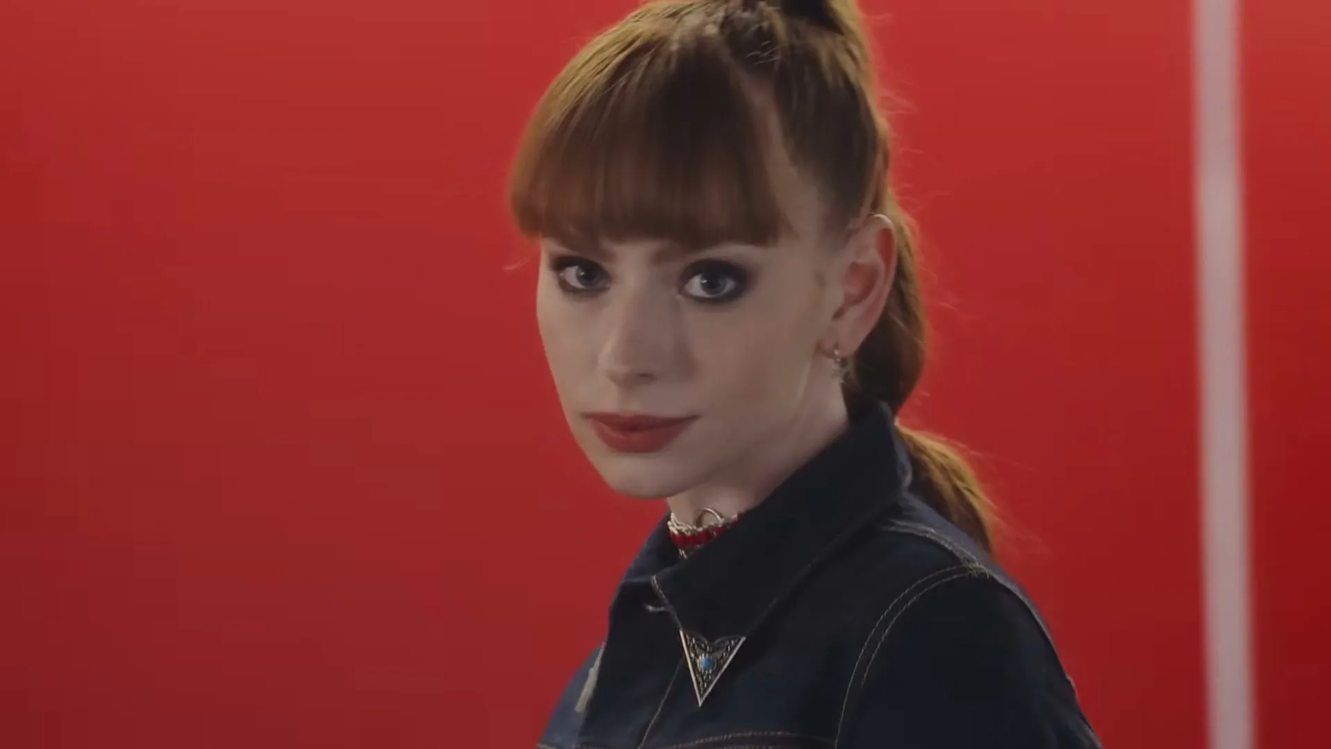 A young woman with red hair and a denim jacket looks over her shoulder with a smirk against a red background.