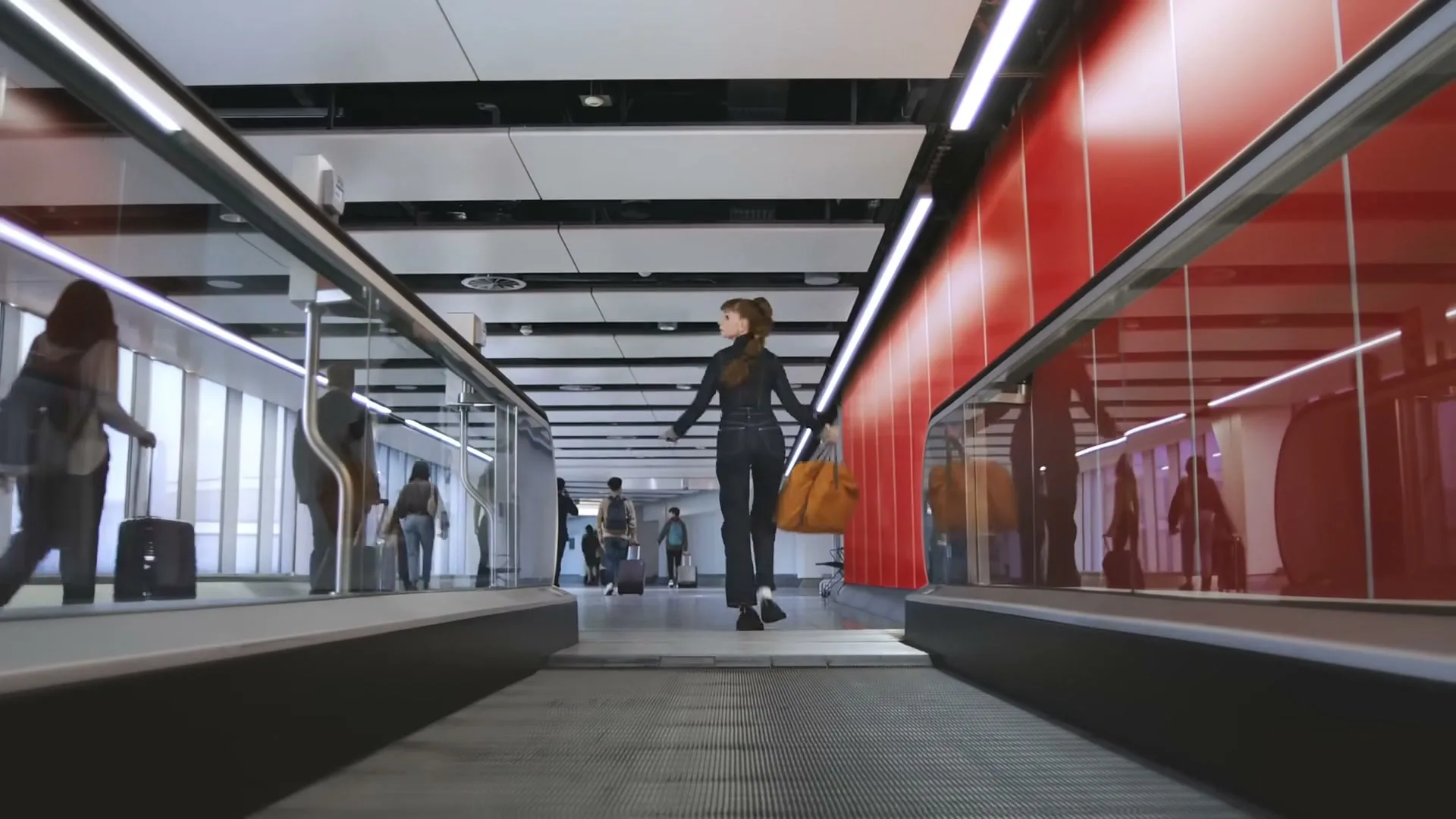 A young woman with auburn hair in a black jumpsuit and yellow bag runs on a moving walkway in a modern airport terminal.