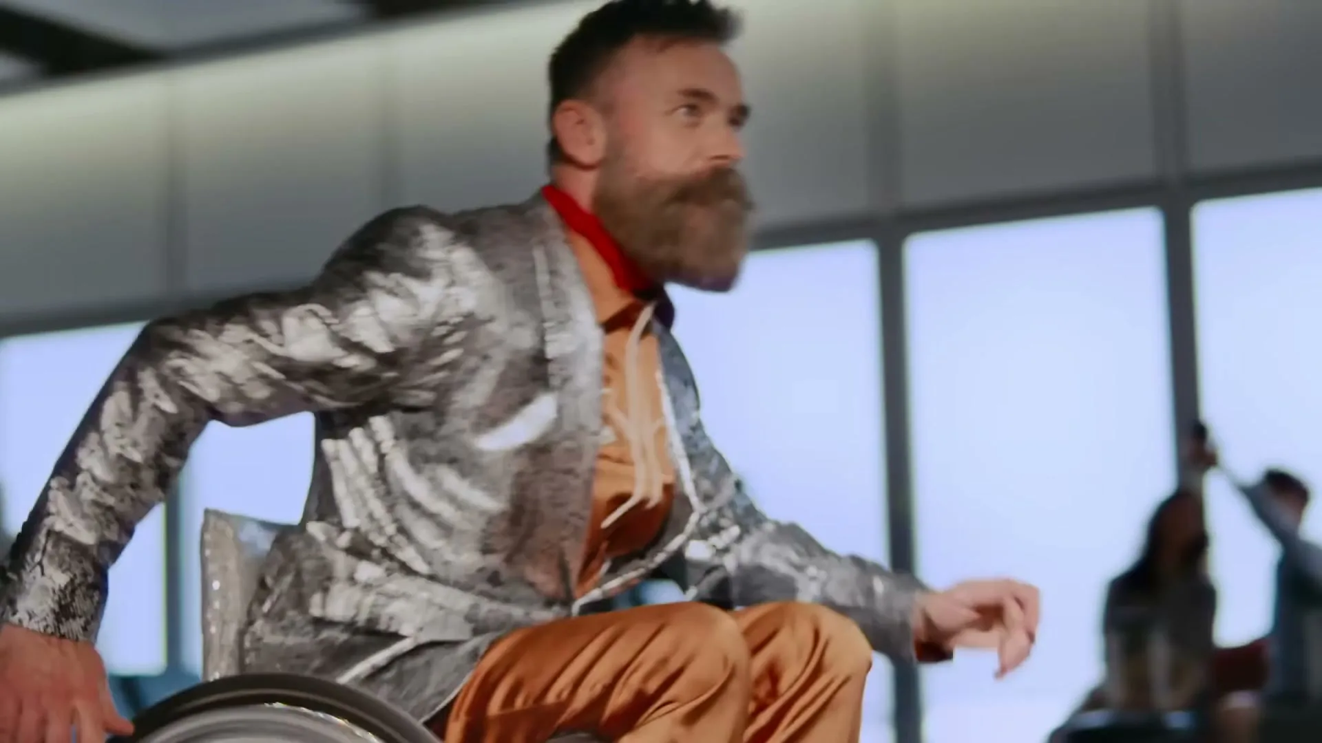 A man with a beard in a wheelchair, wearing a shiny suit, moves across a bright, modern interior space with large windows.