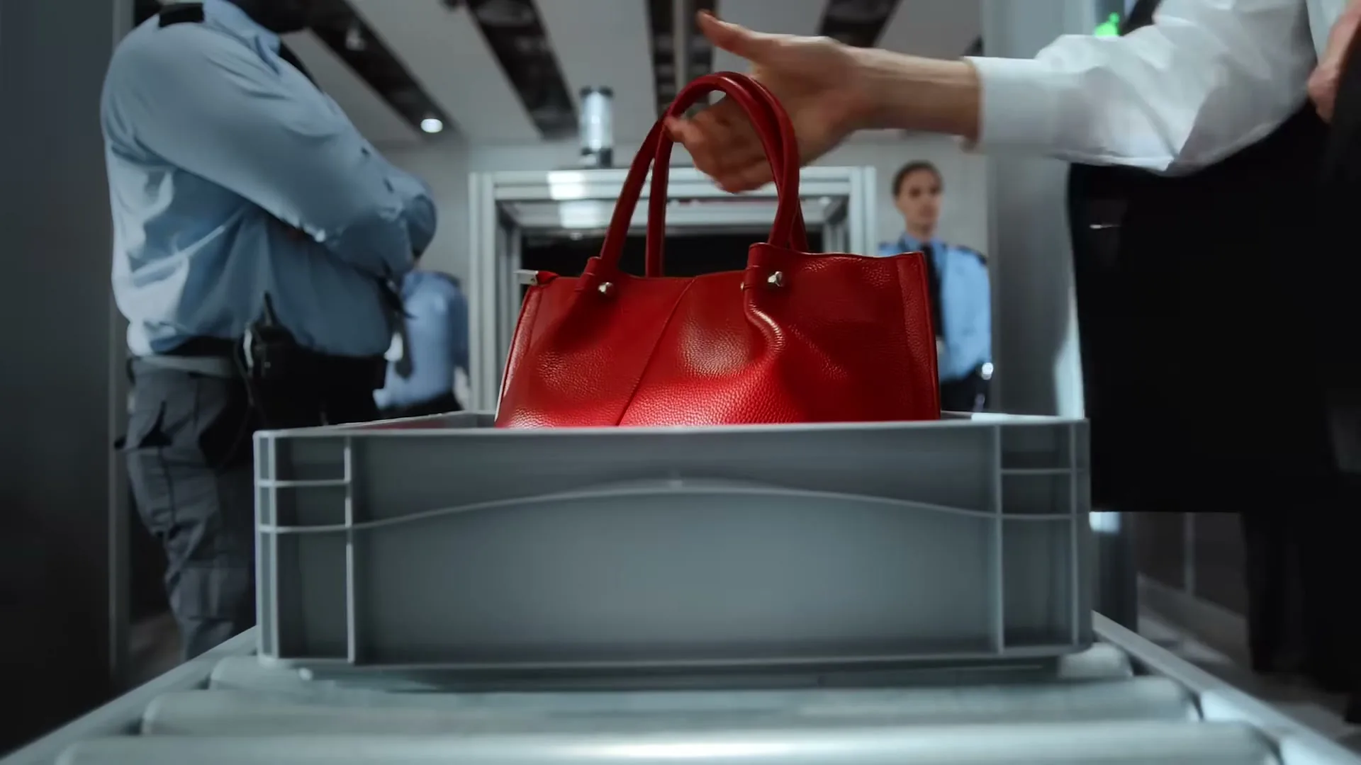 A red handbag is placed into a grey bin on an airport security conveyor belt, with security personnel in the background.