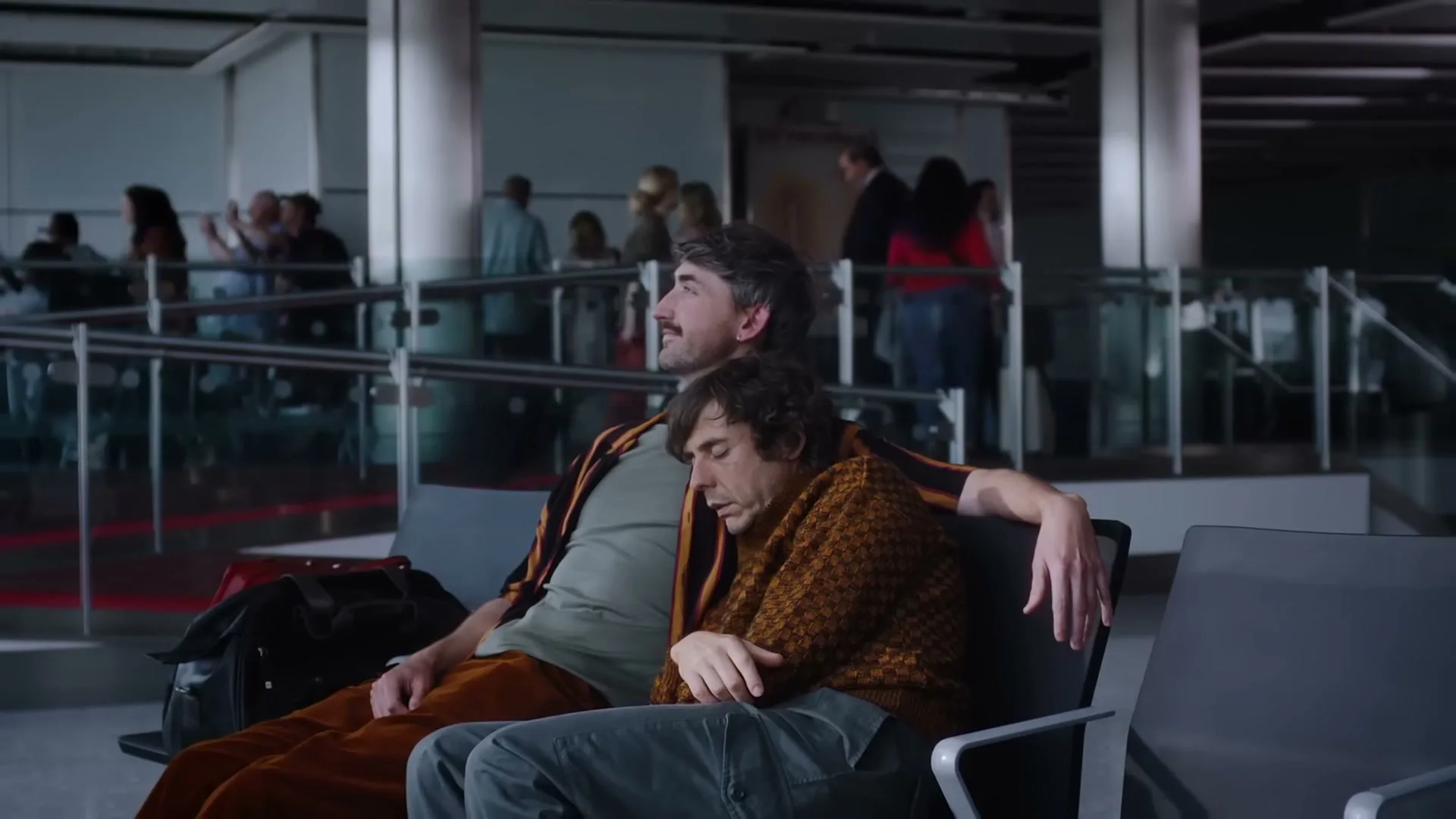 A man with a mustache sits on an airport bench with another man sleeping on his shoulder, in a busy airport terminal.
