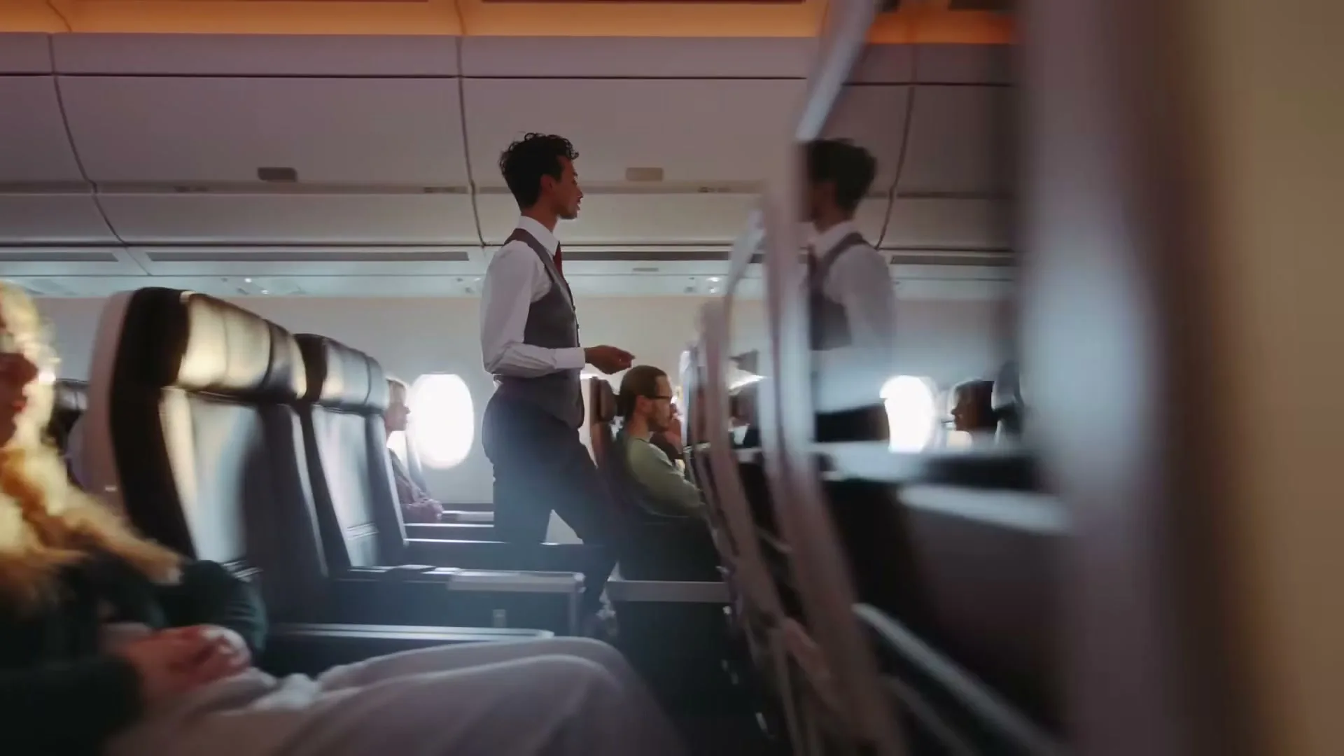 A flight attendant walks down the aisle of an airplane cabin, past passengers seated in their seats, with sunlight streaming through the windows.