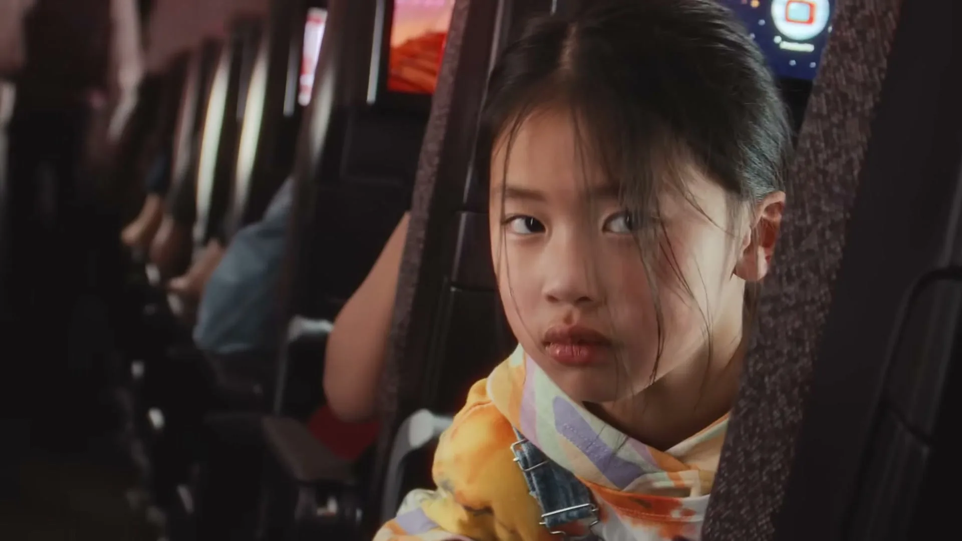 A young girl with dark hair and a colorful hoodie peeks from behind an airplane seat, looking directly at the camera with a curious expression.