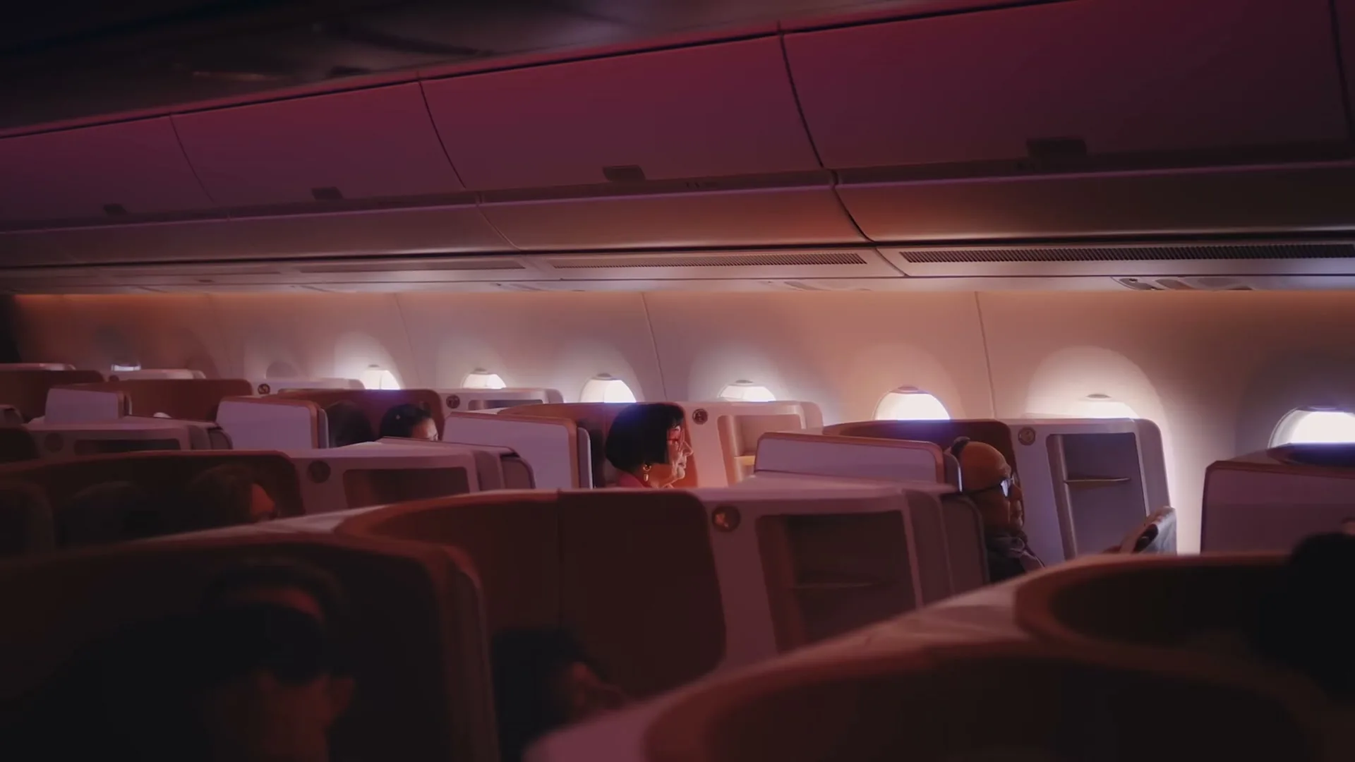 A woman with dark hair and a red top sits in a business class airplane cabin, looking out the window.