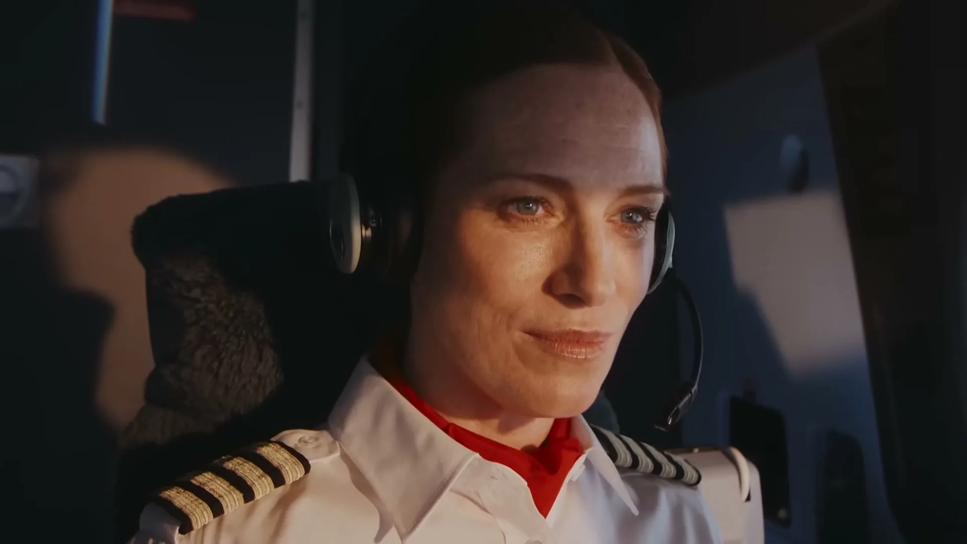 A female pilot in uniform and headset looks forward with a slight smile in the cockpit of an airplane.
