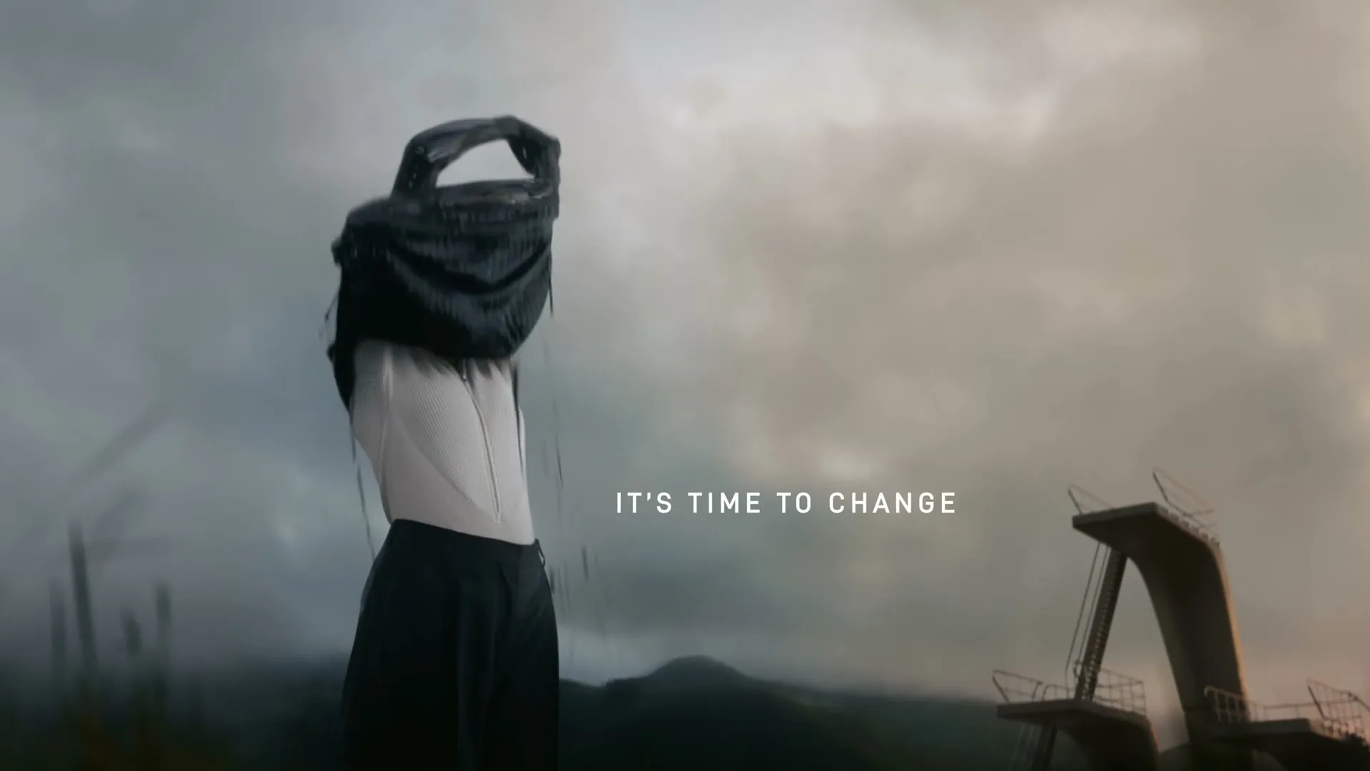 A woman covered in black goo sheds her outer layer to reveal clean clothes, standing in a desolate, cloudy landscape with a diving board structure.