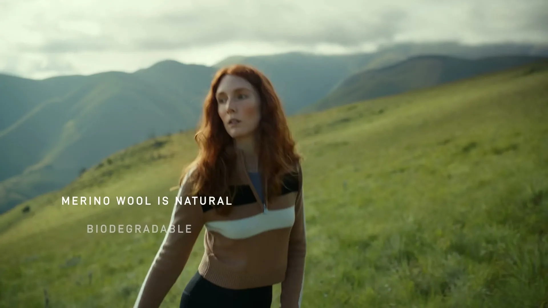 A young woman with long red hair, wearing a brown, white, and black striped top, walks through a grassy mountain landscape.