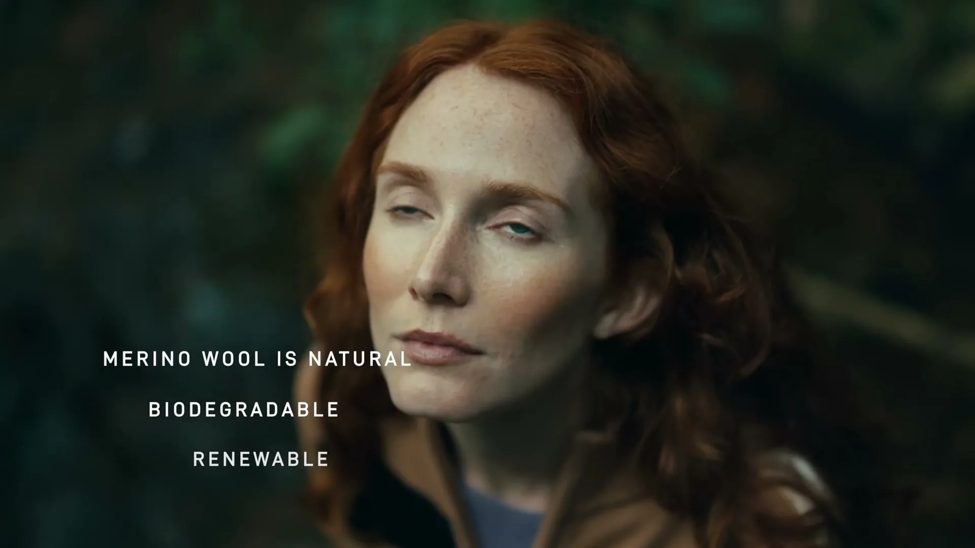 A woman with red hair and blue eyes looks up, then down, then up again, in a forest.