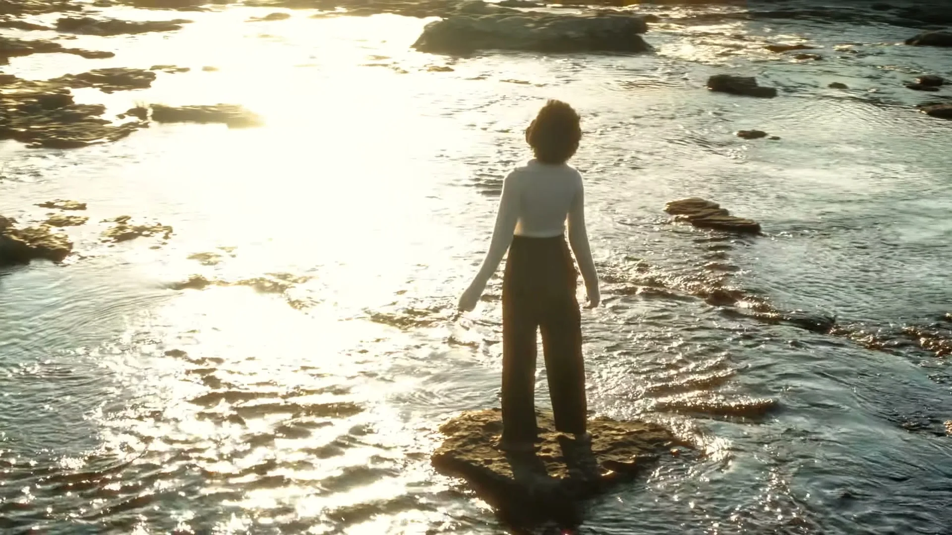 A woman with dark curly hair stands on a rock in a shallow river, looking towards the bright setting sun, in a wide shot.