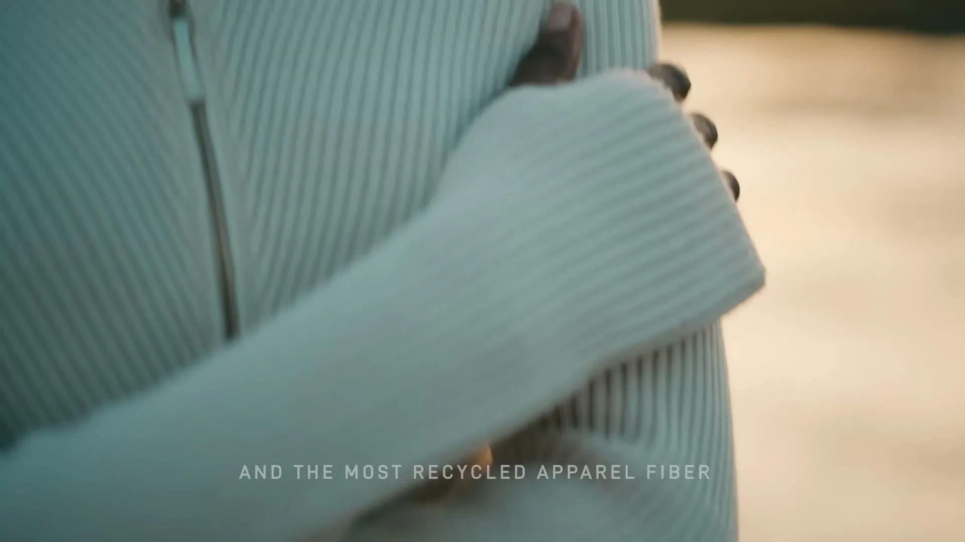 A person in a white ribbed sweater with a zipper is seen from the chest up, with their hand gently touching their arm.