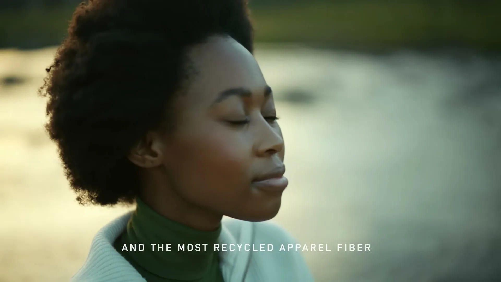 A young Black woman with curly hair, wearing a green turtleneck and white sweater, stands with her eyes closed by a body of water at golden hour.