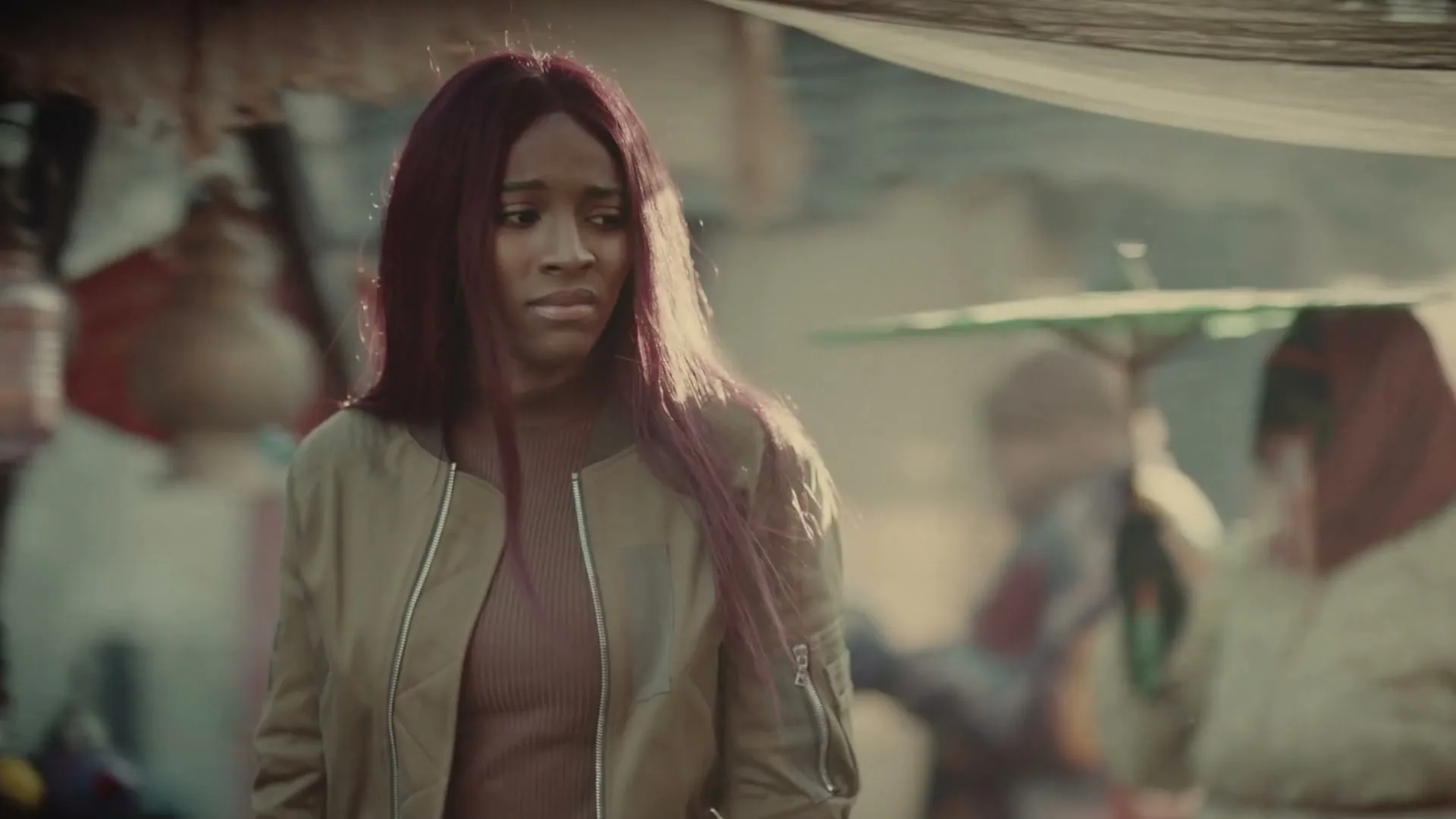 A young woman with long red hair and a bomber jacket looks around a dusty, crowded marketplace with a worried expression.