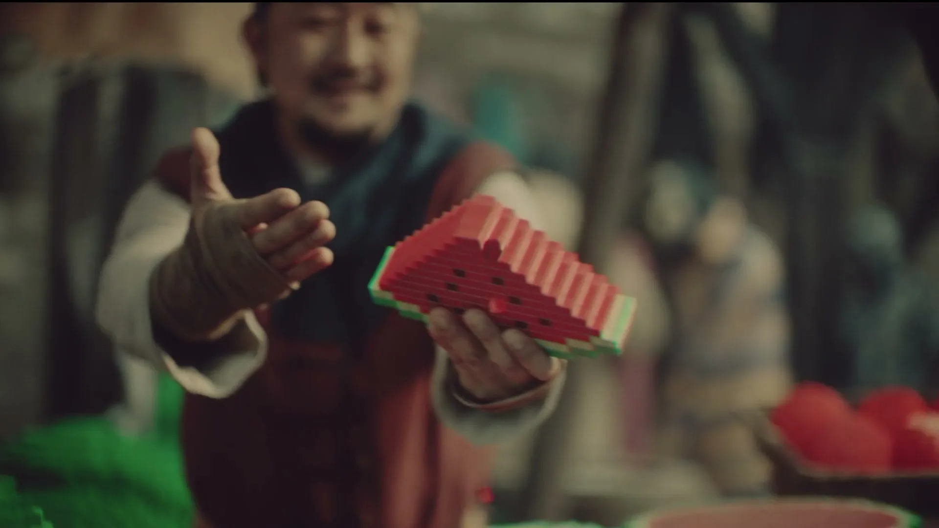 A smiling male vendor in traditional attire holds out a pixelated watermelon slice at a bustling market stall.