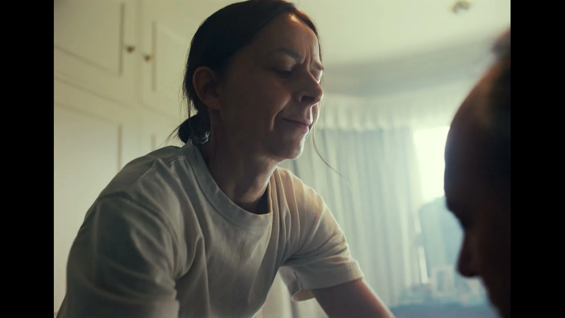 A woman in her 30s, wearing a white t-shirt, looks distressed as she speaks to someone off-camera in a brightly lit room.