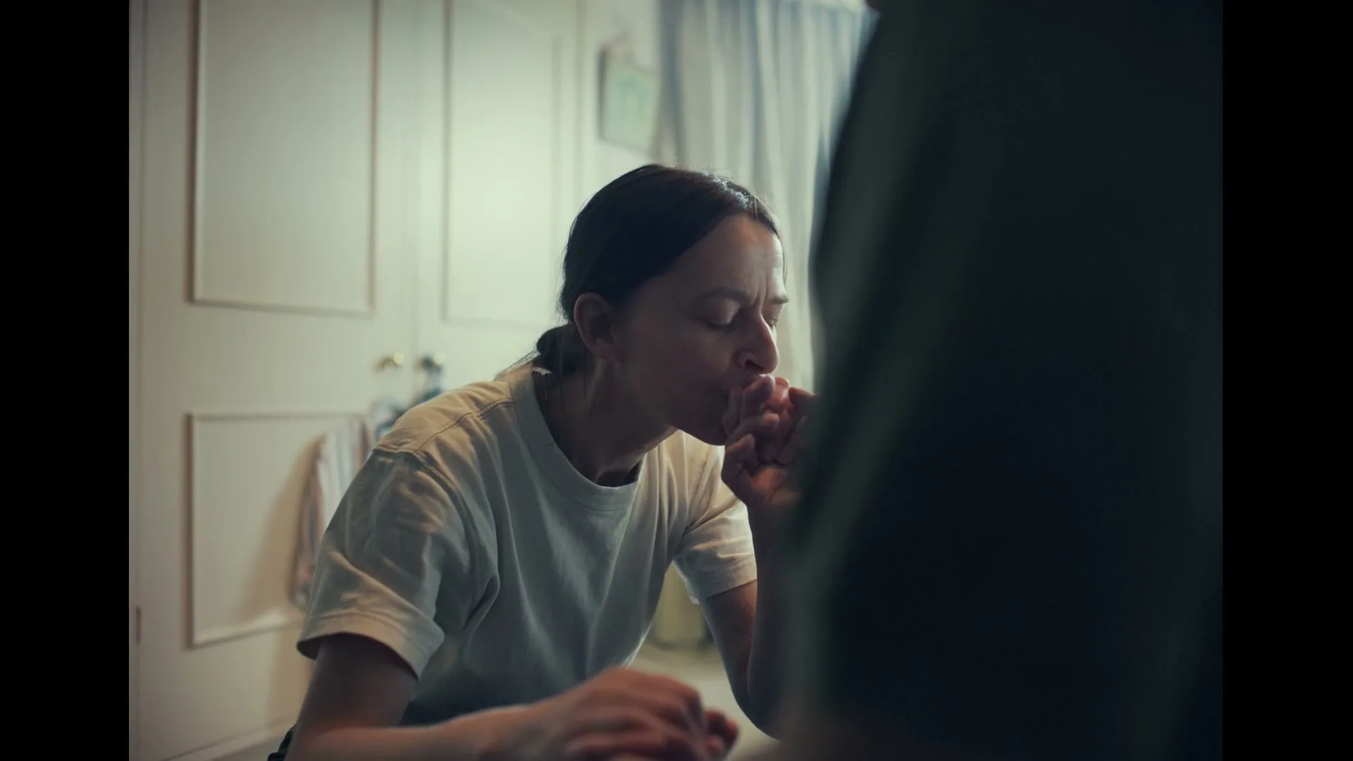 A woman with dark hair in a white t-shirt smiles and then kisses the hand of an unseen person in a softly lit room.