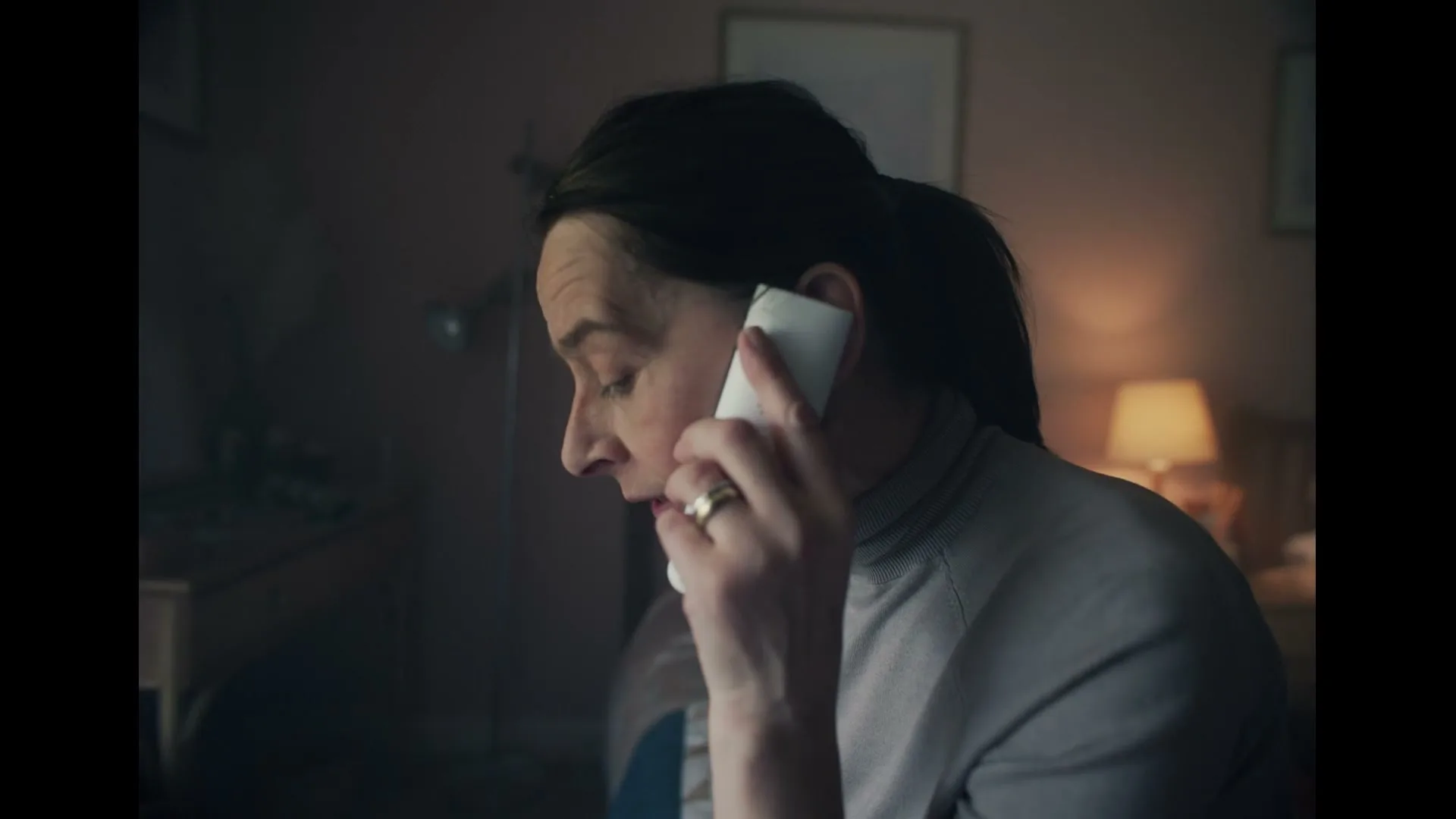 A woman with dark hair in a ponytail, wearing a grey turtleneck, holds a white phone to her ear, looking downcast in a dimly lit room.