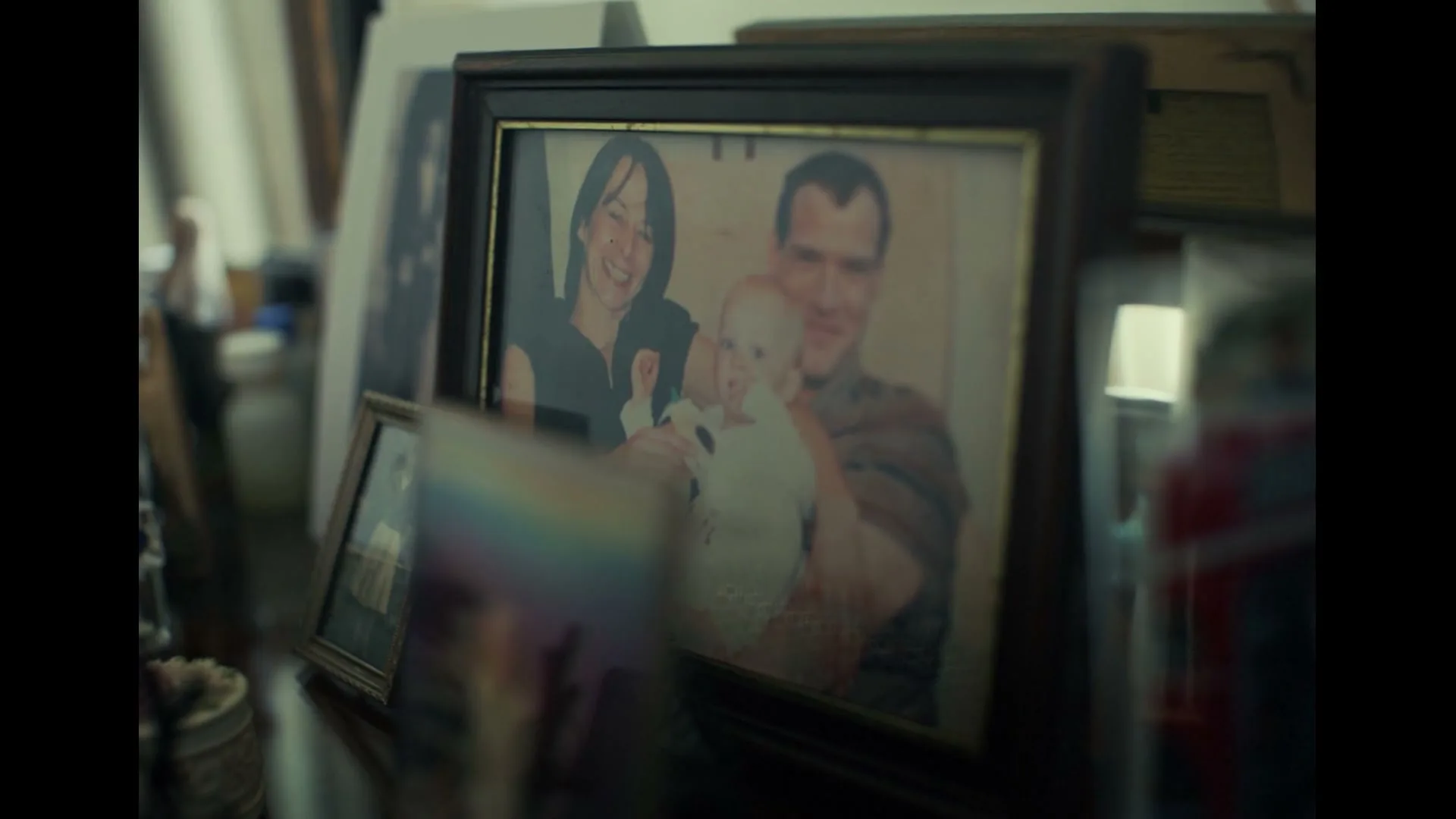 A framed photo of a smiling family, a man and woman holding a baby, sits on a cluttered surface in an interior setting.