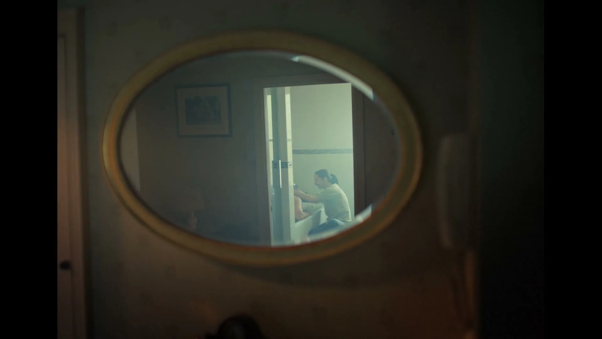 A woman in her 30s, wearing a green shirt, is seen reflected in an oval mirror, hunched over in a bathroom.