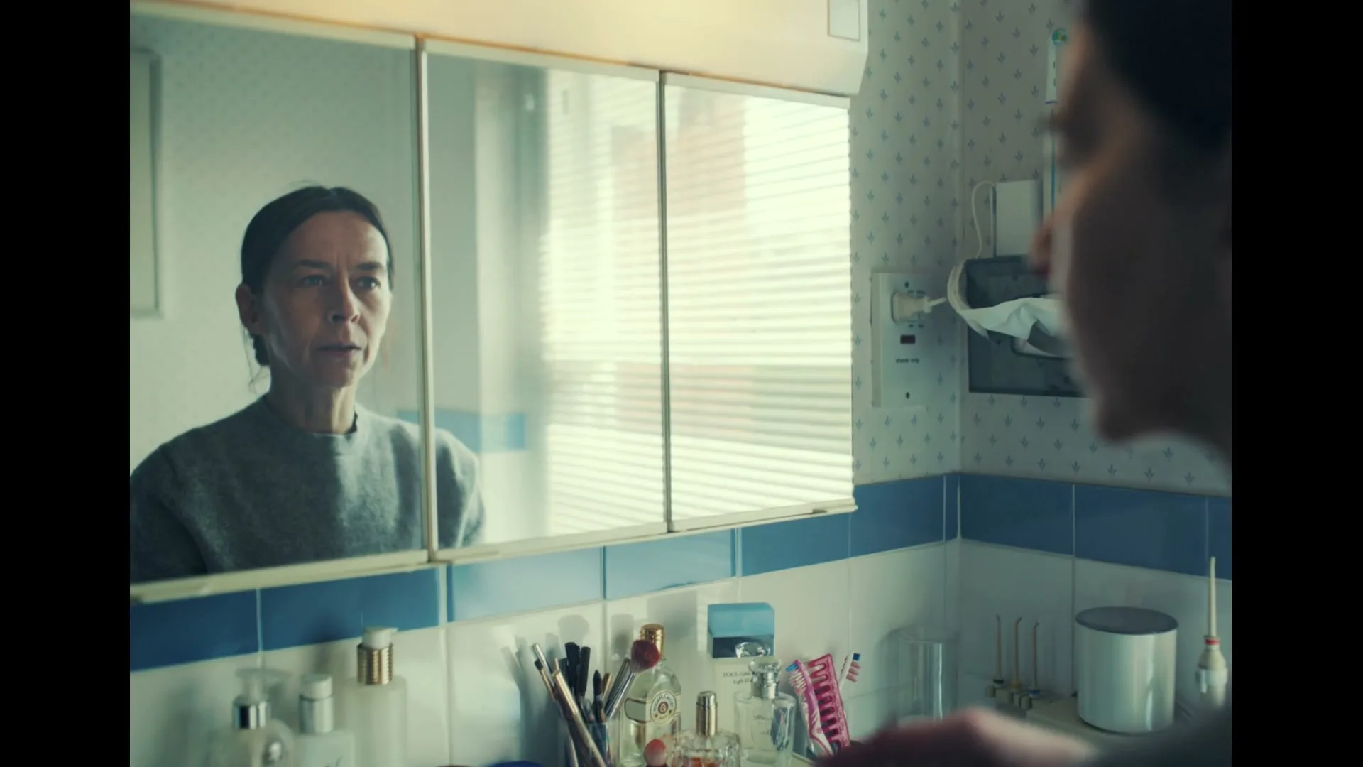 A woman with dark hair in a grey sweater looks at her reflection in a bathroom mirror, touching her face with a concerned expression.