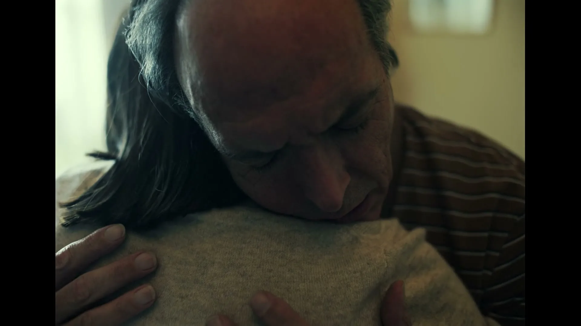 A man with thinning hair and a striped shirt hugs a woman, resting his head on her shoulder, in a softly lit interior.