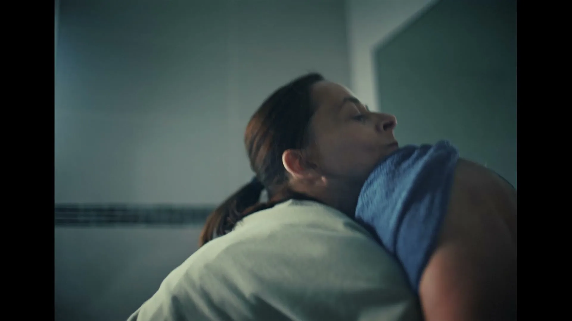 A woman with dark hair in a ponytail hugs another person, burying her face into their shoulder, in a dimly lit room.