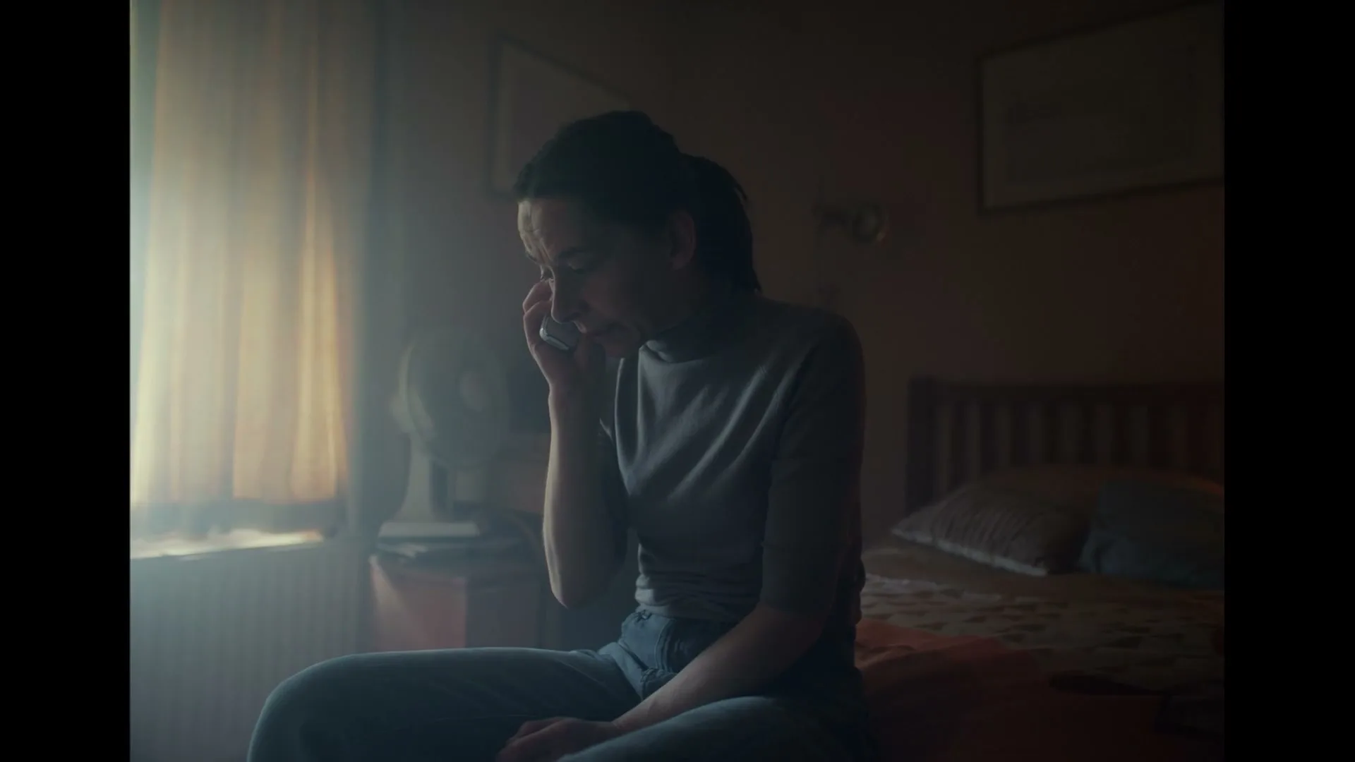 A woman in her 30s sits on a bed, holding a phone to her ear, looking distressed in a dimly lit bedroom.